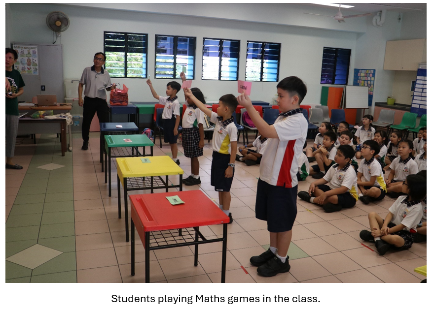 Children in school uniforms hold up pink cards near colorful tables while classmates sit on the floor watching.