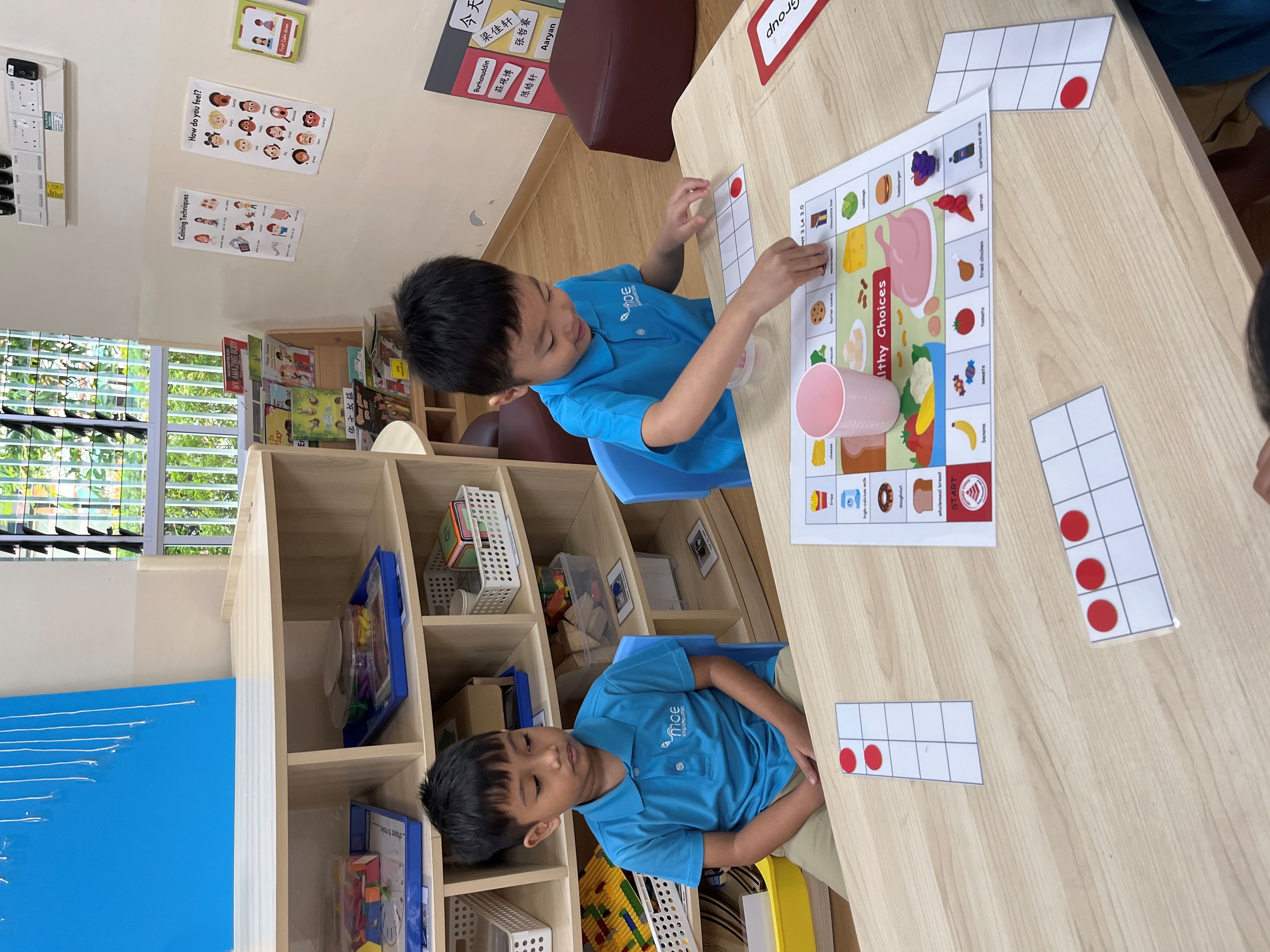 Two young boys in blue uniforms sit at a table playing a Healthy Choices board game with counting cards.