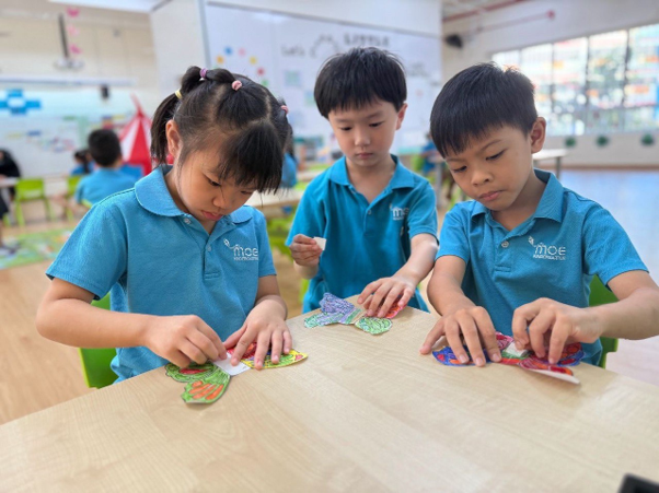 Three young children in blue uniforms sit at a table doing a craft activity with colorful cut-out pieces in a classroom.