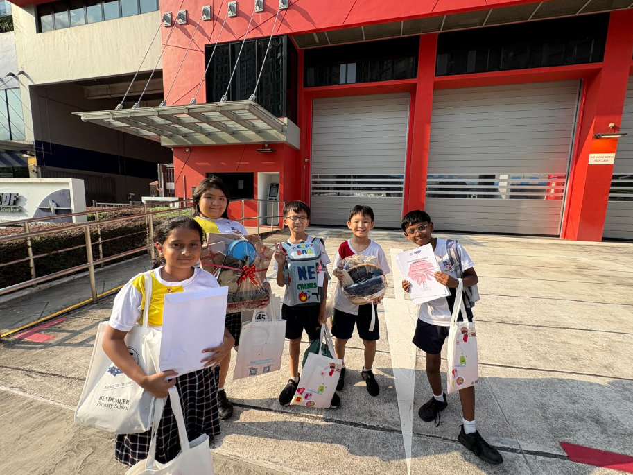 Five school children stand outdoors in front of fire station, holding bags, gift baskets, and papers.
