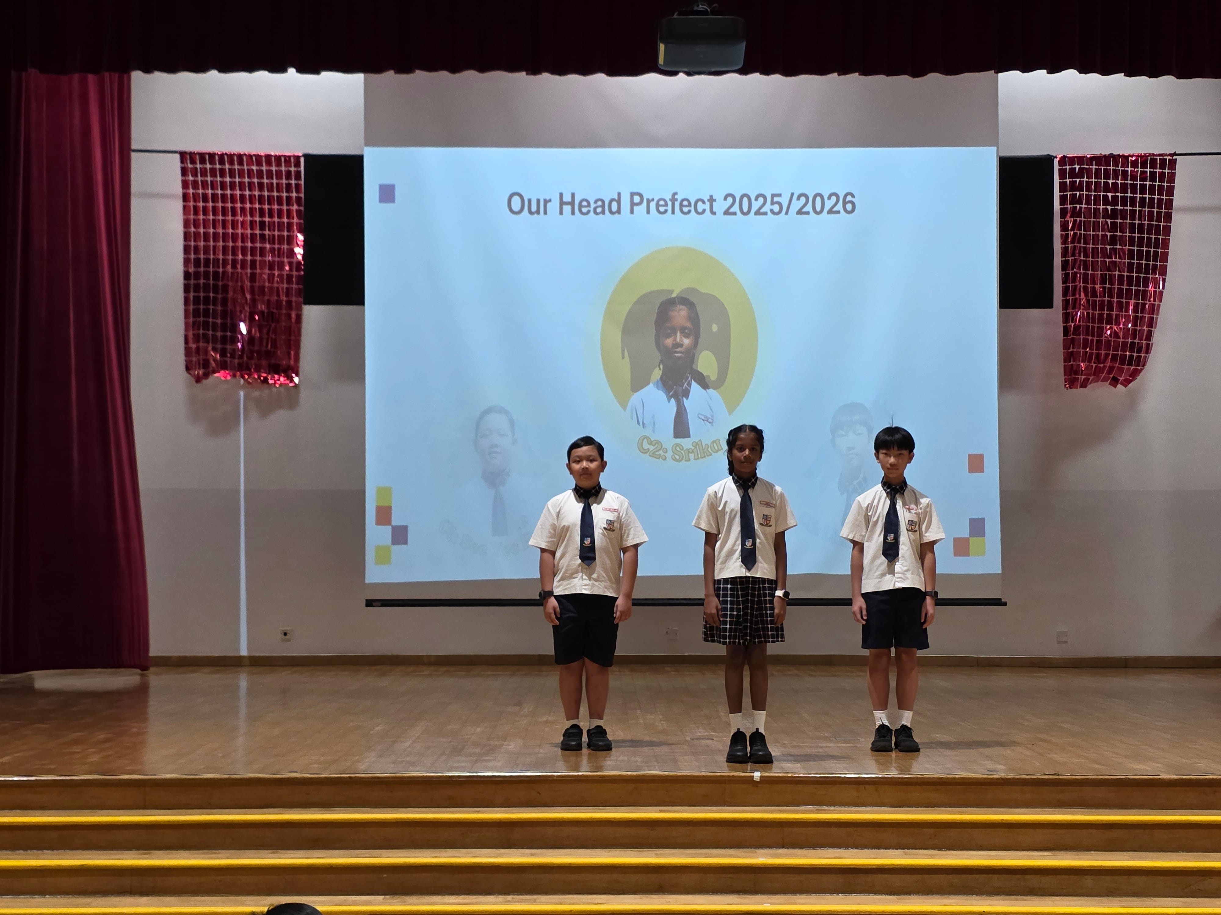 Three school students in uniforms stand on a stage before a projected slide reading "Our Head Prefect 2025/2026."