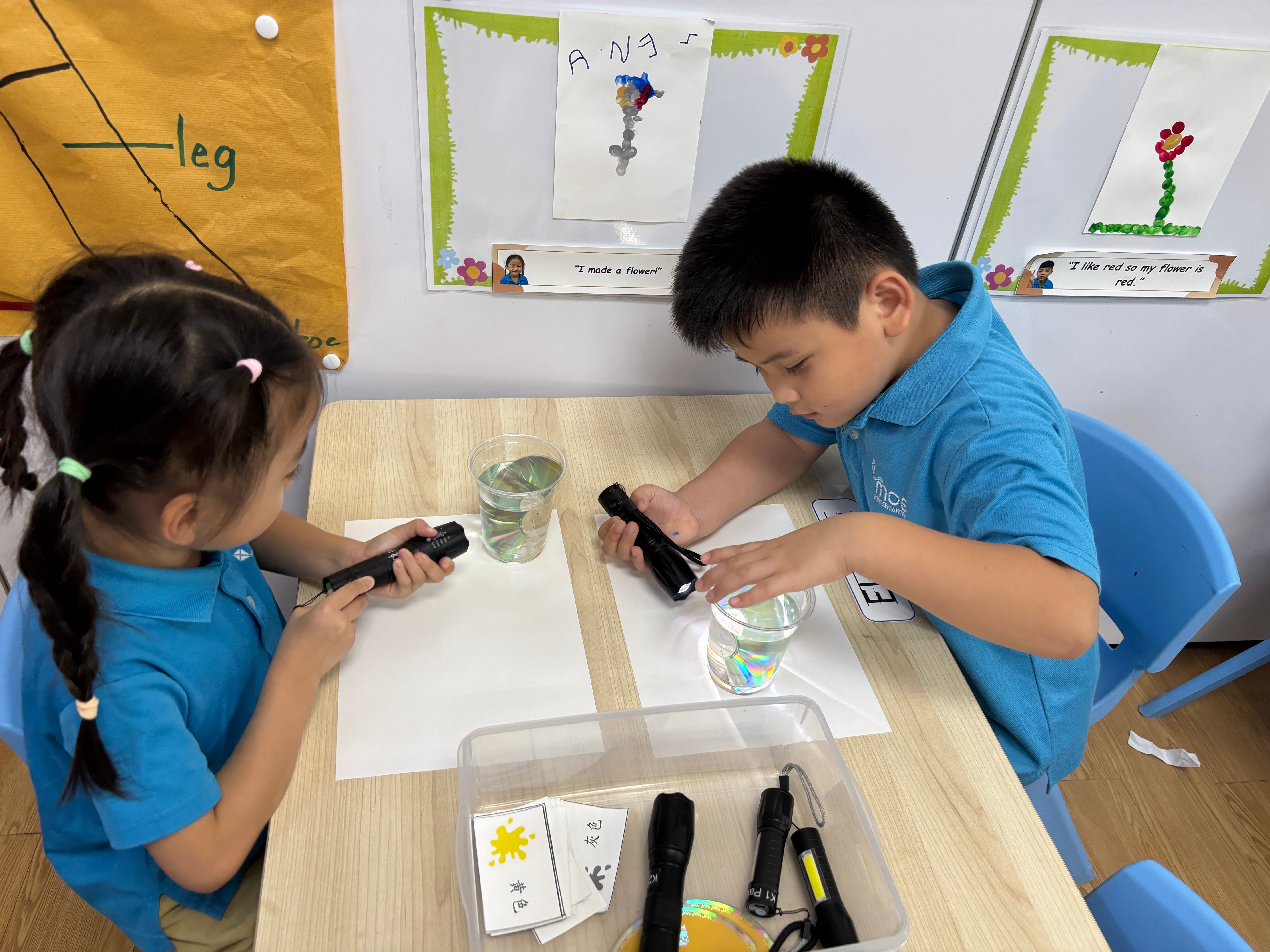 Two children at a table shine flashlights into cups of water over white paper during a classroom activity.