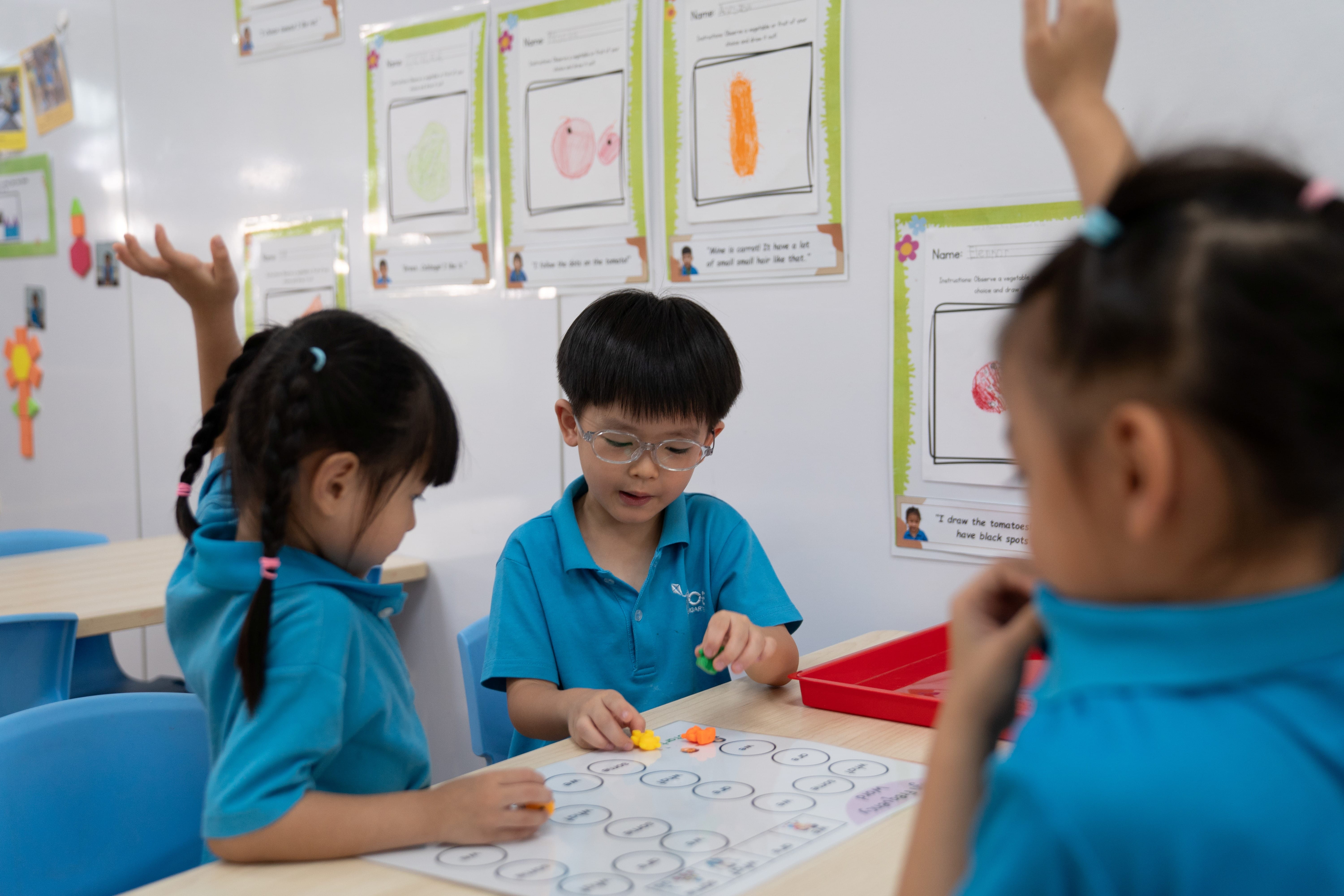 Children in blue uniforms play a board game at a classroom table, with artwork displayed on the wall.
