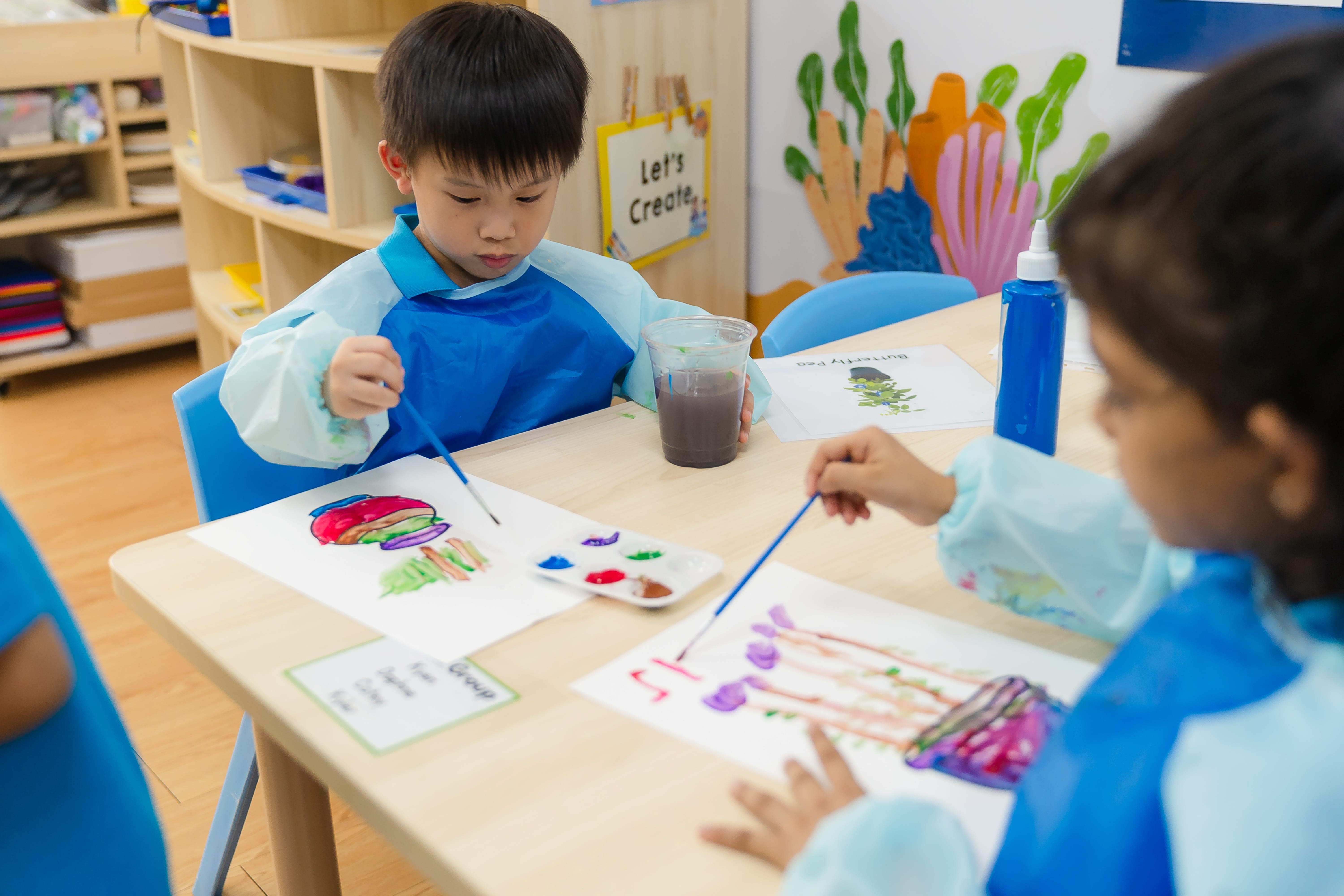 Two children in blue smocks paint pictures at a classroom table with brushes, paints, and a cup of water.