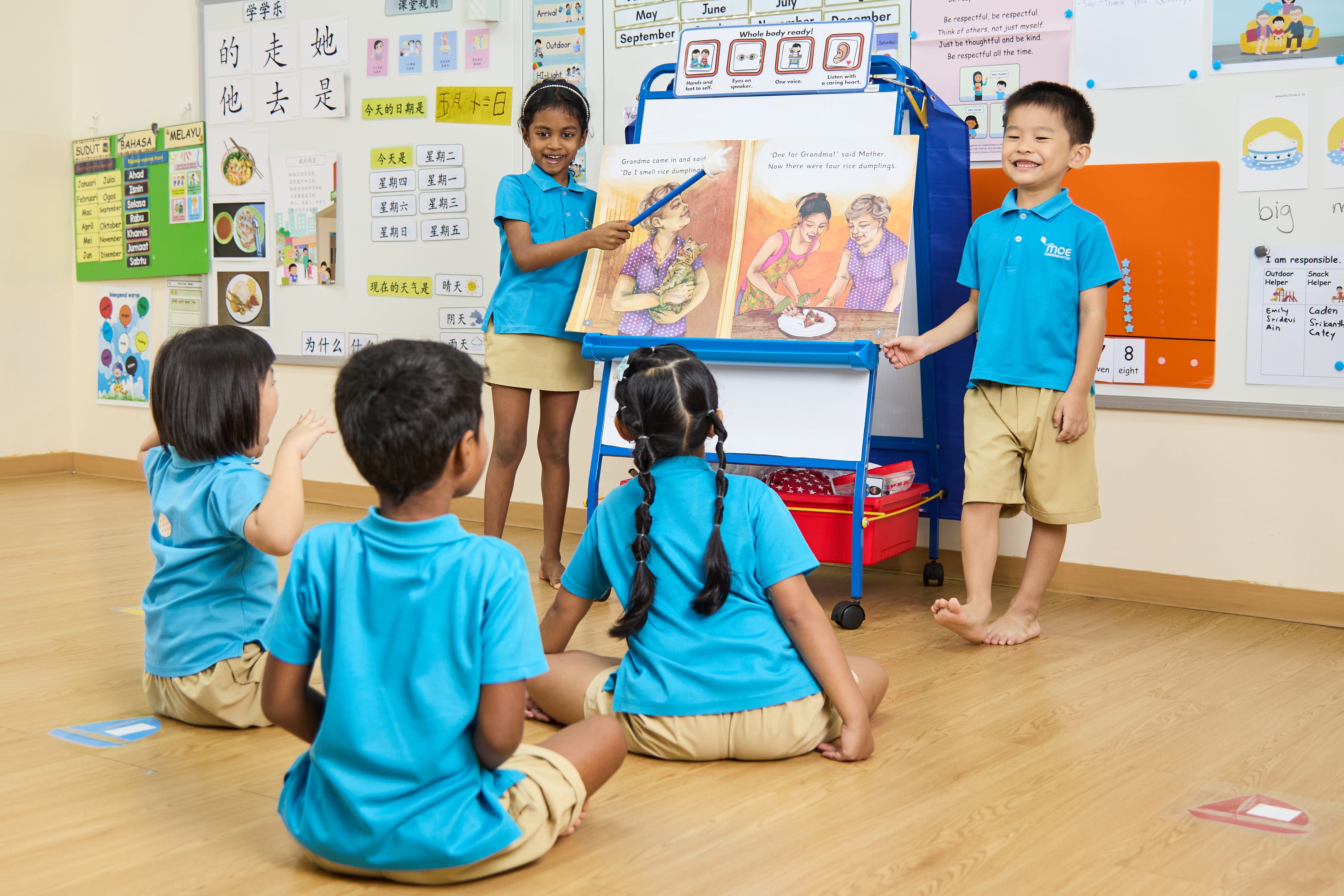 Two children stand by a large open picture book on an easel while classmates sit on the floor listening.