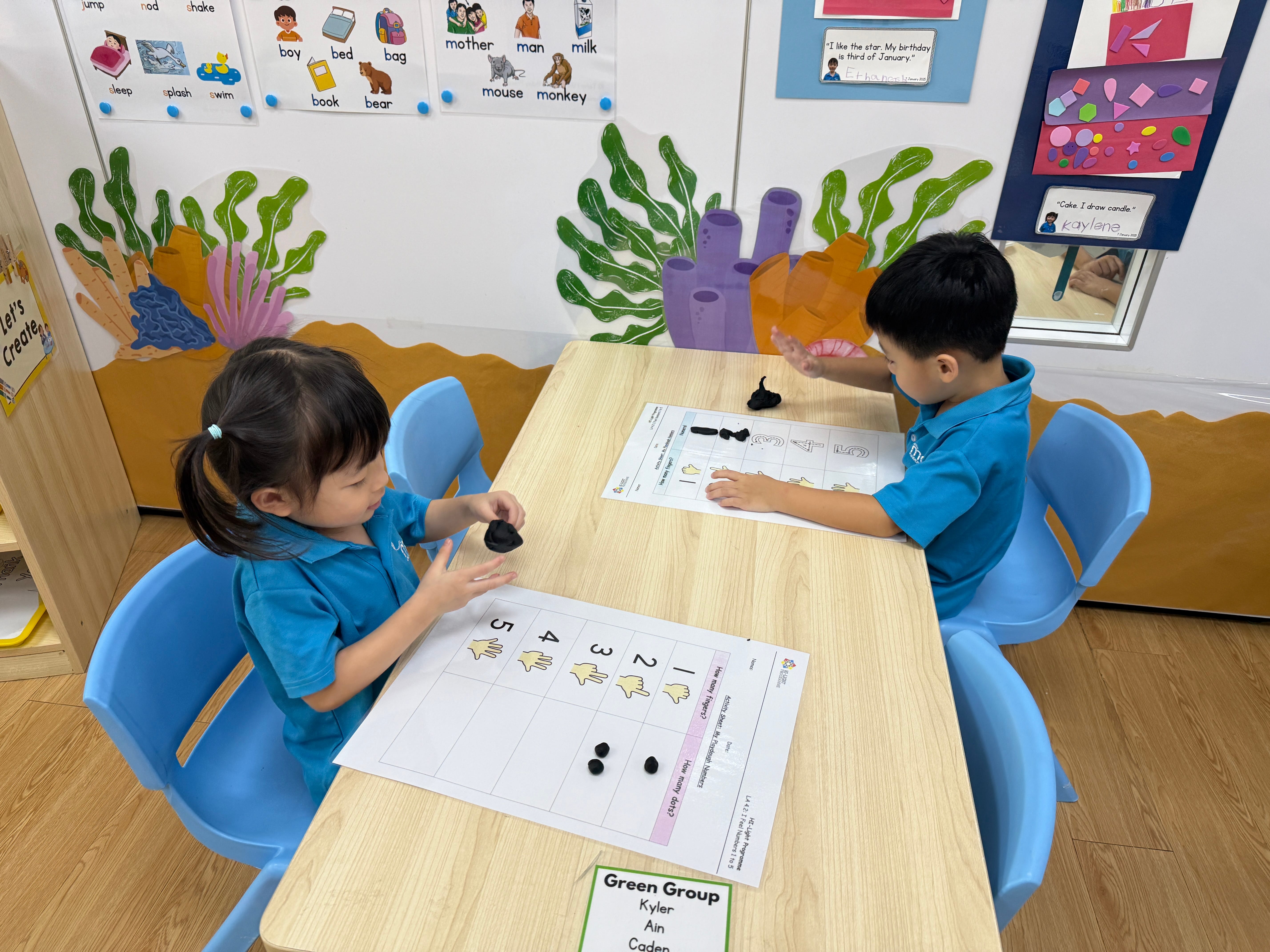 Two children in blue shirts sit at a table, using black clay pieces on number worksheets.