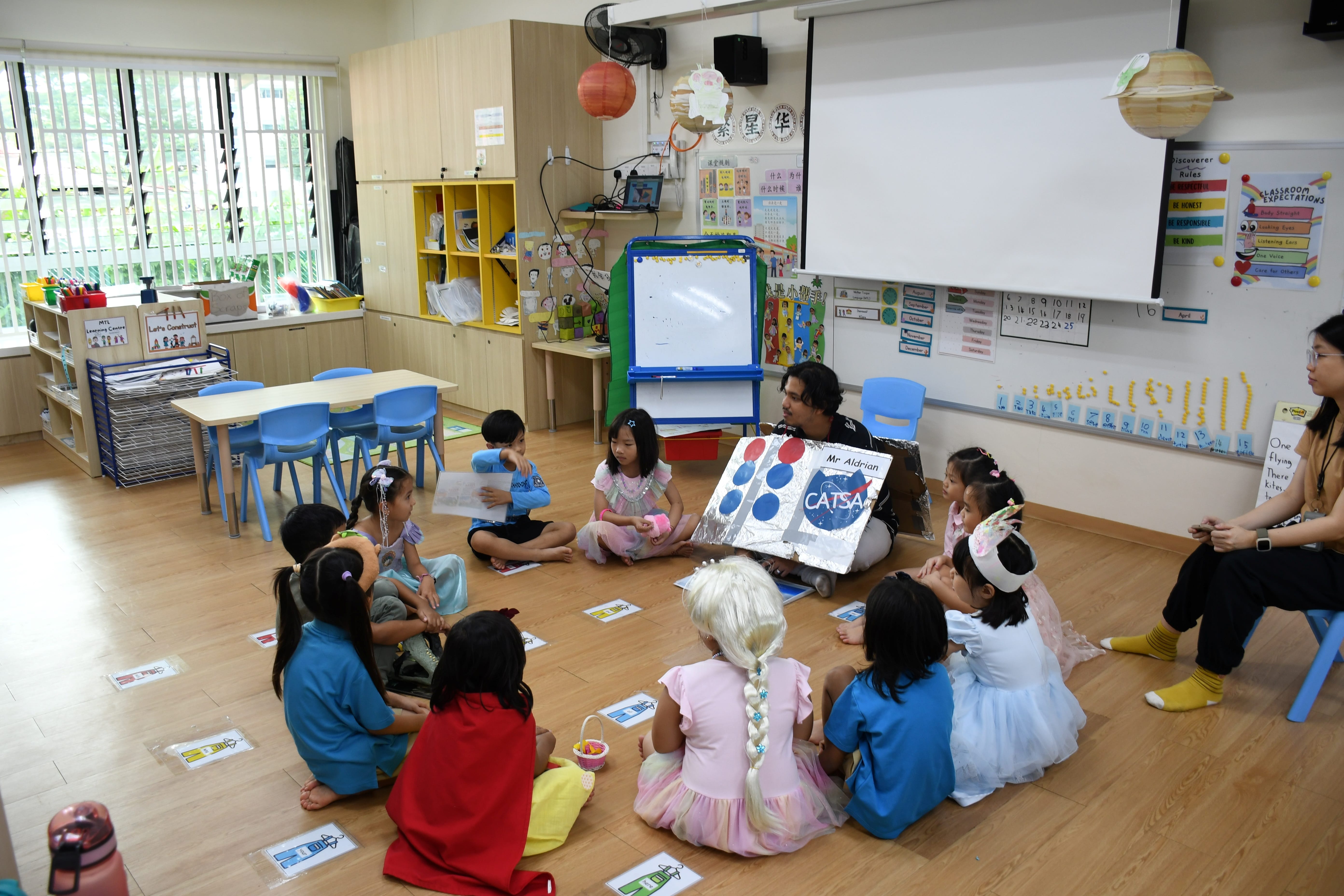 Teacher shows a CATSA sign as children in costumes sit in a circle on a classroom floor.
