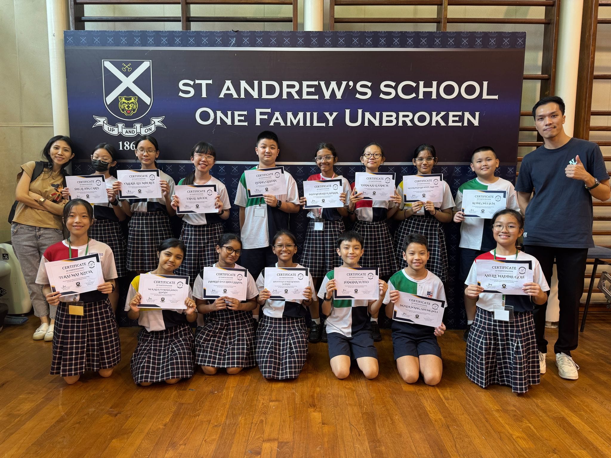 Students and teachers pose holding certificates in front of a St Andrew's School One Family Unbroken banner.