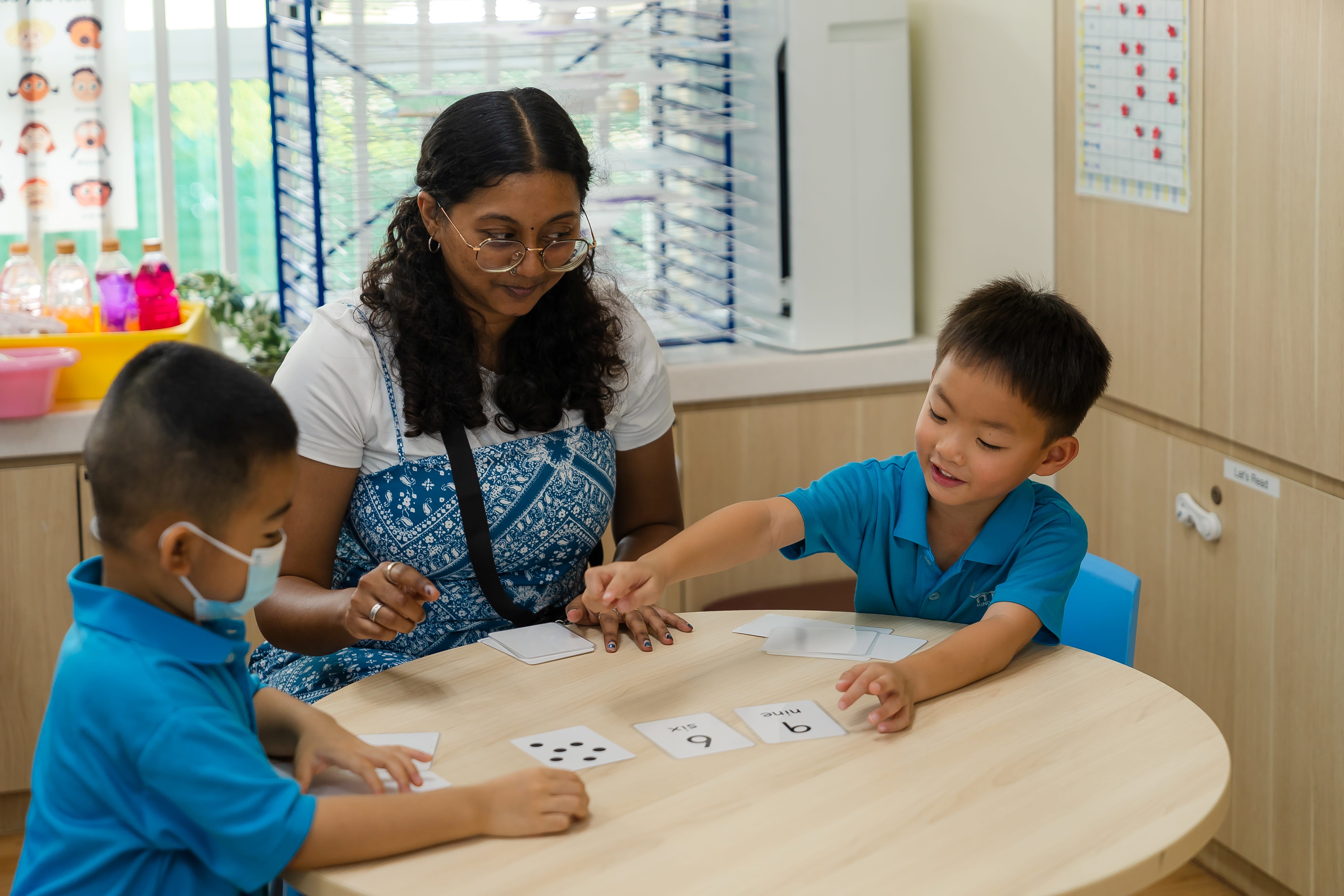 Teacher with two children at a round table, using number and dot flashcards in a classroom.