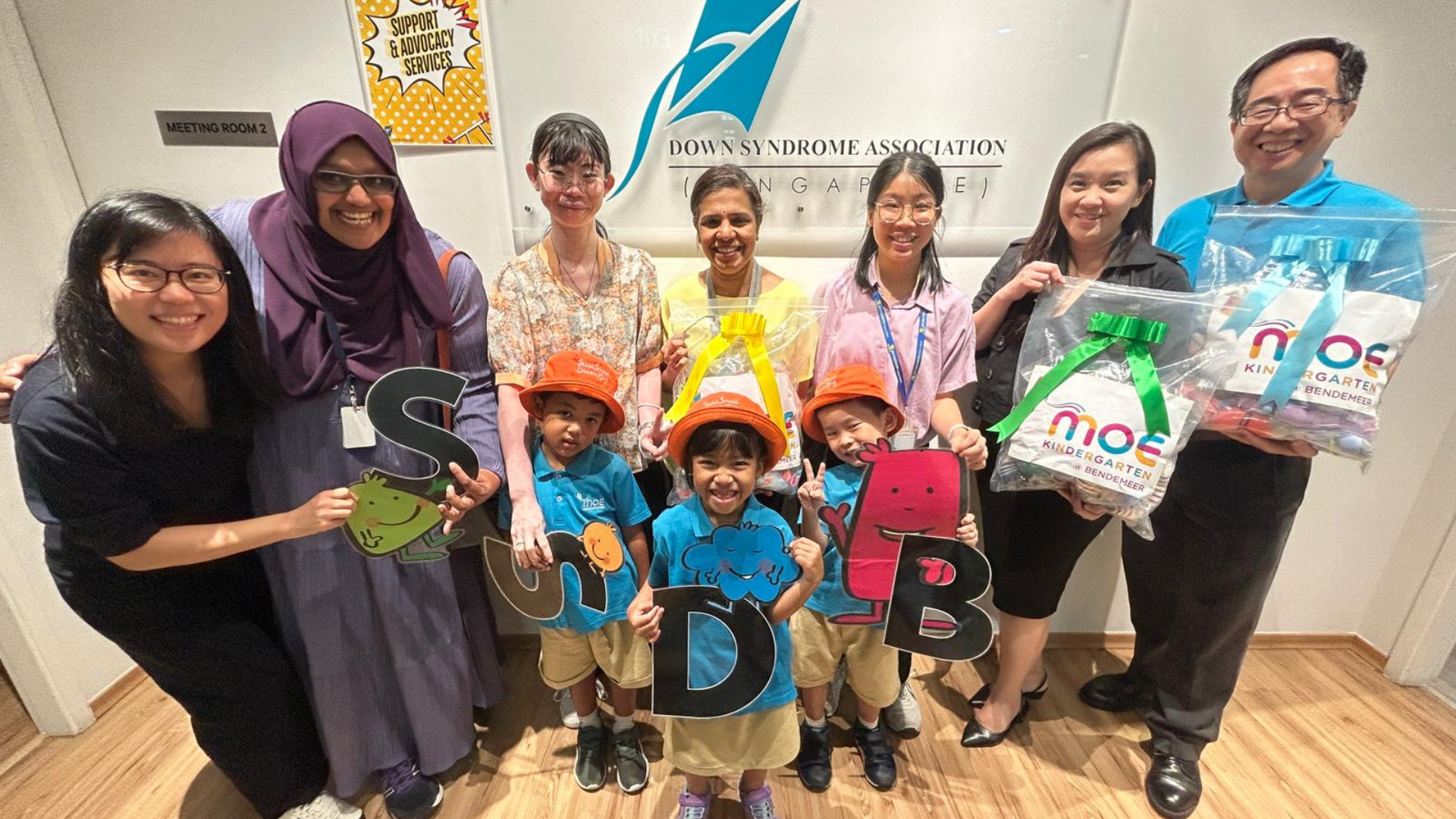 Adults and children in MOE Kindergarten uniforms pose holding letters spelling SUDB at Down Syndrome Association Singapore.