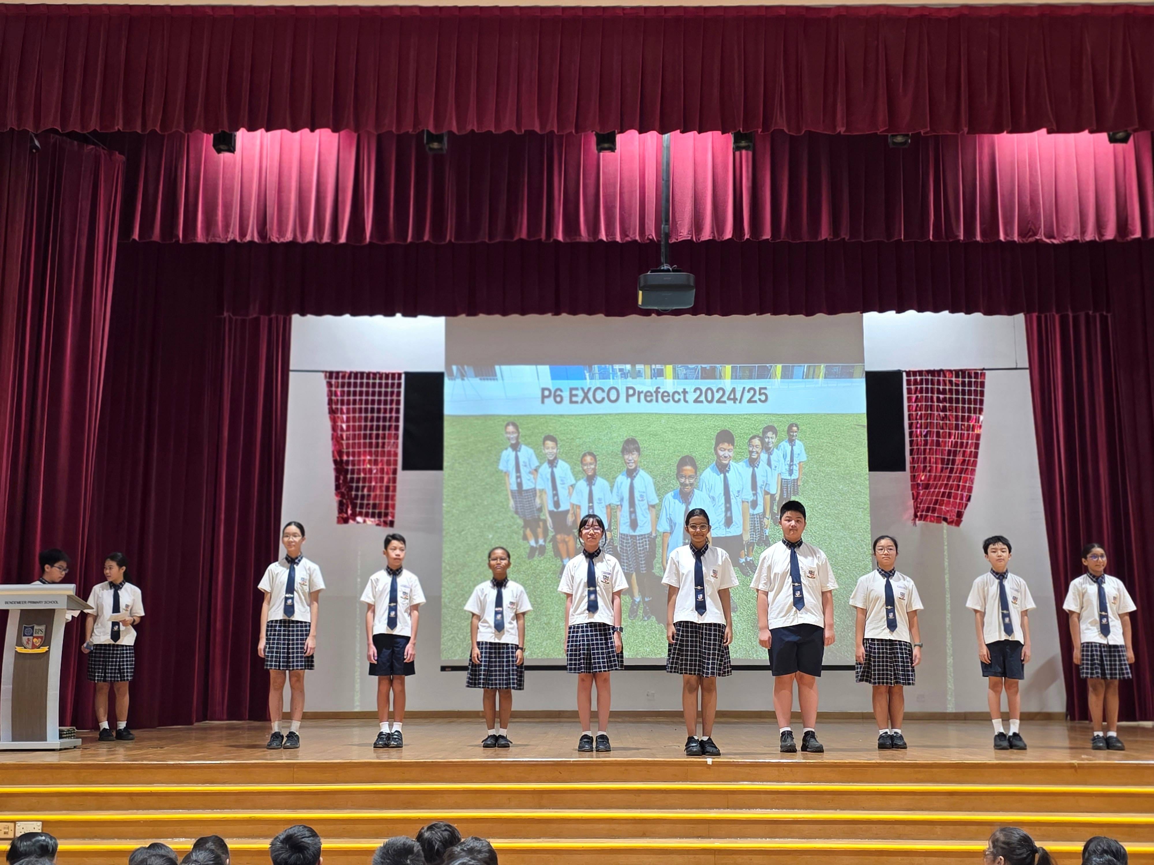 Students in school uniforms stand on a stage before a screen reading "P6 EXCO Prefect 2024/25."