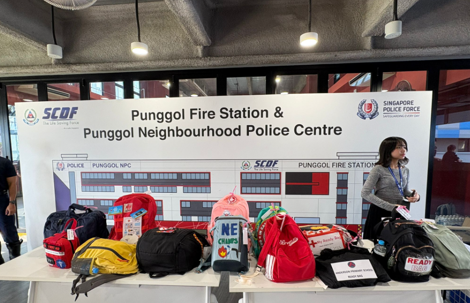 A table with various bags in front of a Punggol Fire Station and Neighbourhood Police Centre banner.