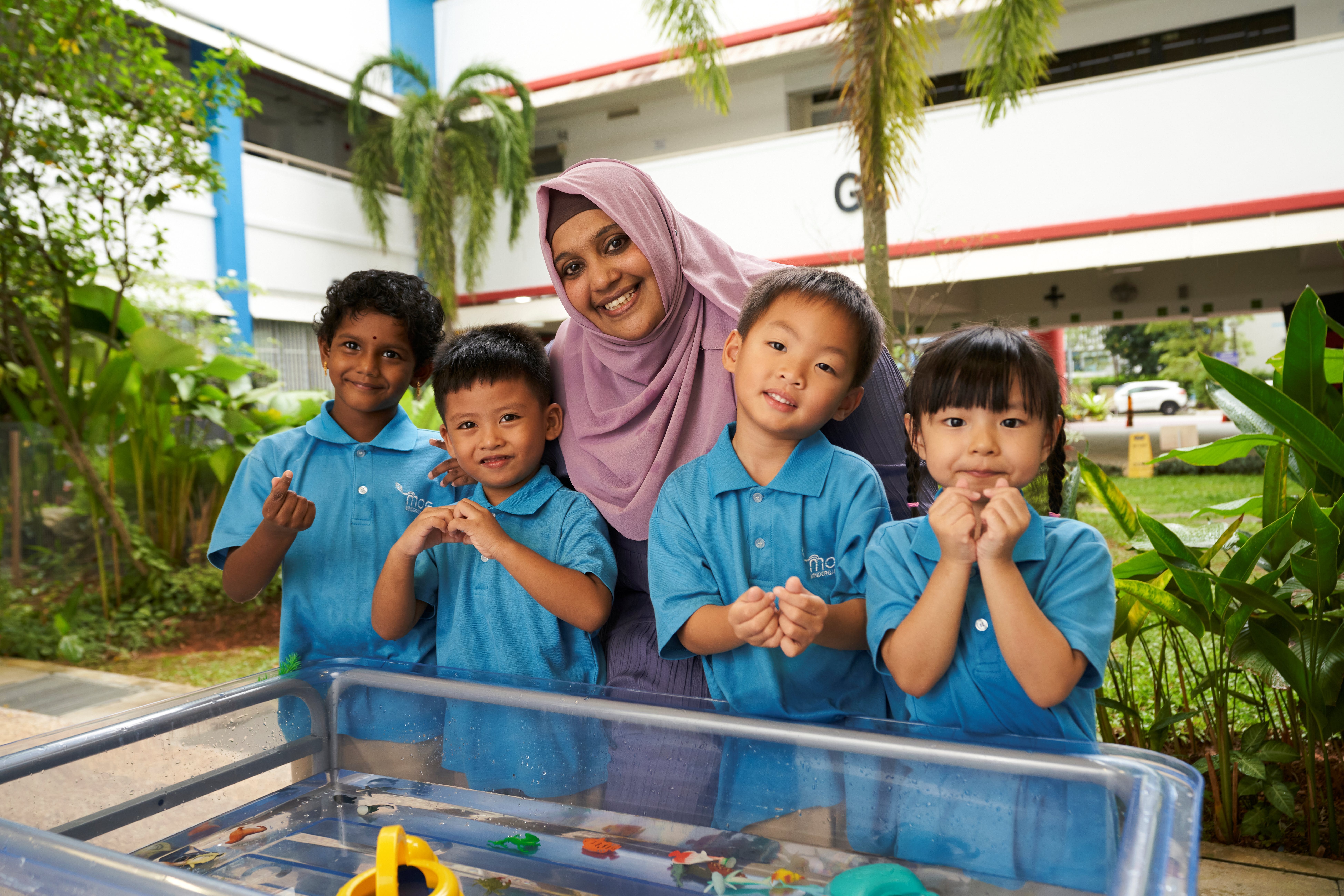 Deputy Centre Head smiles with four children in blue uniforms by a water tray with floating toys outdoors