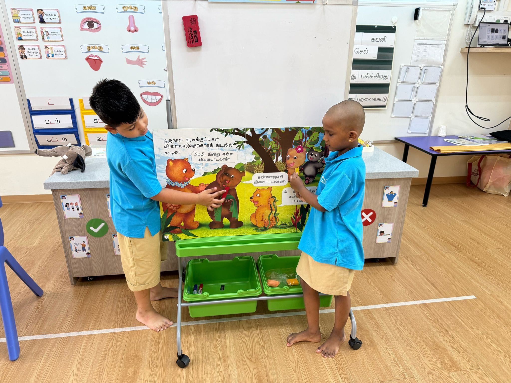 Two young boys in blue uniforms hold a large Tamil storybook with bear illustrations in a classroom.
