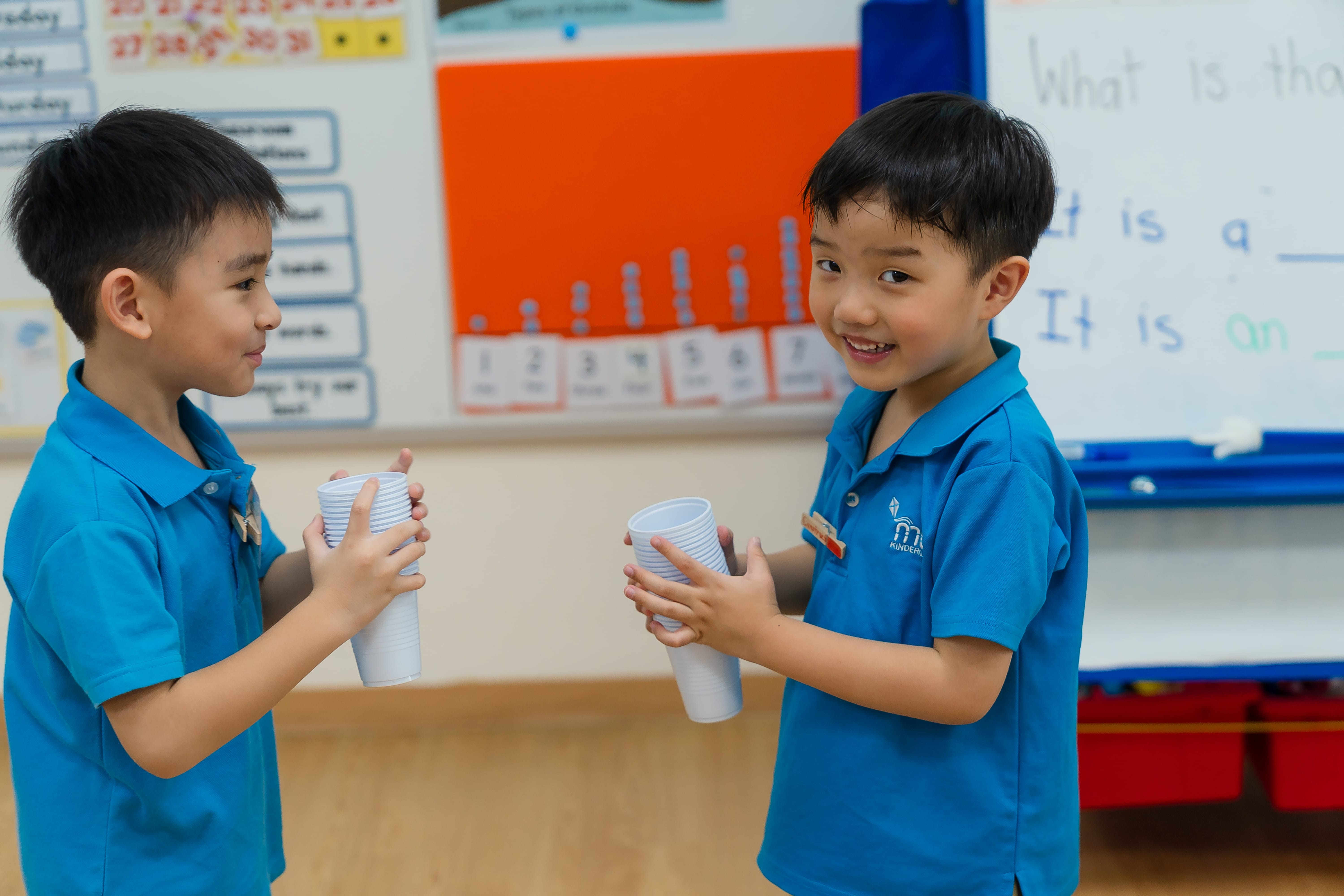 Two boys in blue shirts smile in a classroom, each holding a stack of white plastic cups.