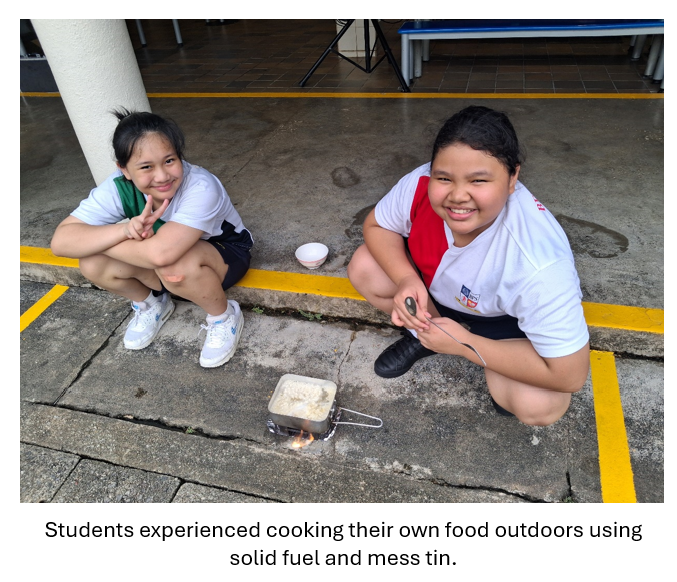 Two smiling girls in white uniforms crouch outdoors beside a small mess tin cooking over a solid fuel flame.