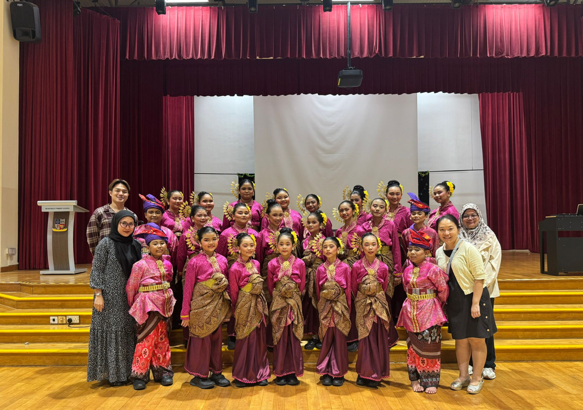 A group of performers in bright pink traditional costumes with gold headpieces pose on a school stage with red curtains.