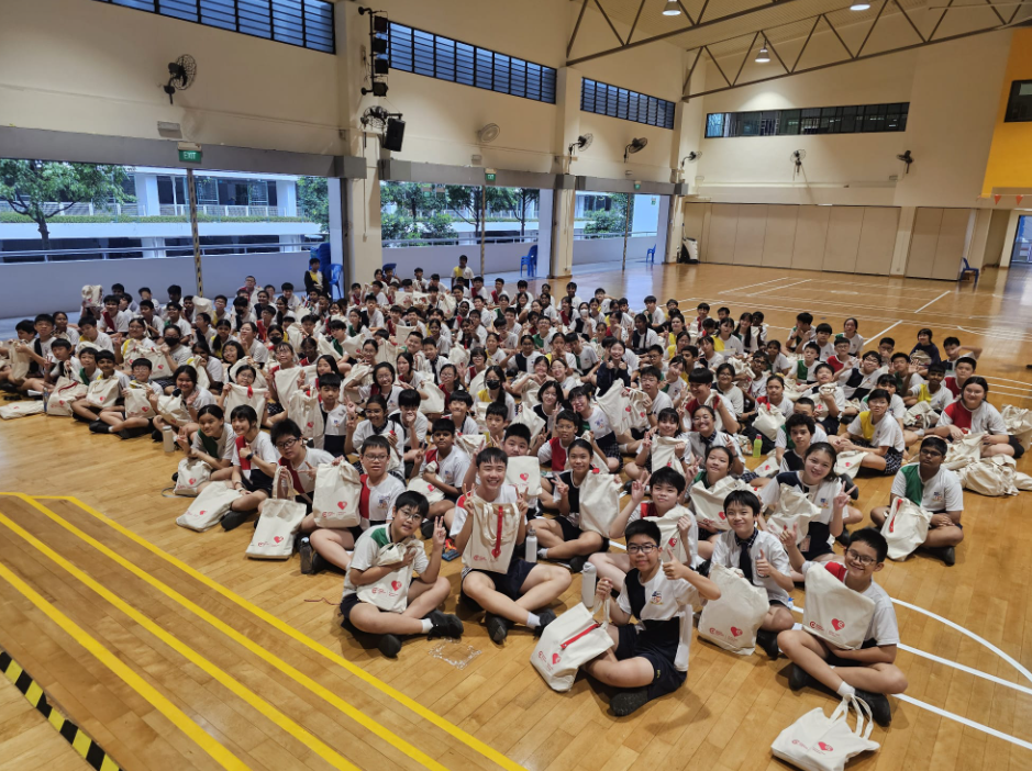 Large group of students in white uniforms sitting on a gymnasium floor, holding white tote bags and smiling.