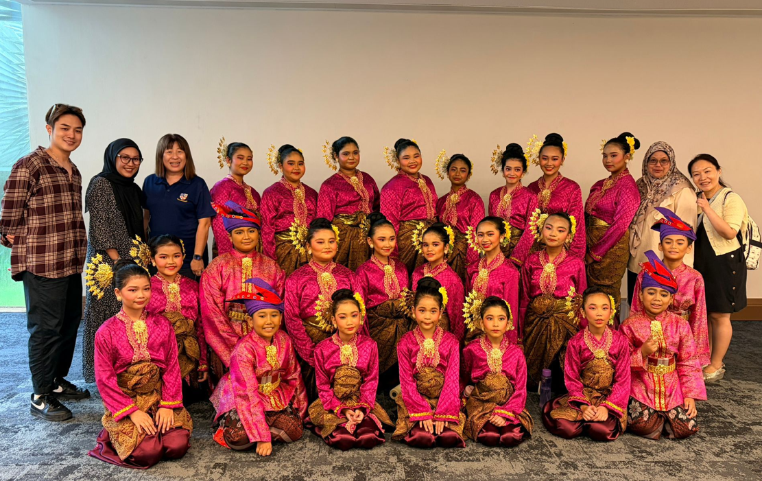 A large group of young dancers in bright pink traditional costumes with gold accessories pose for a group photo.