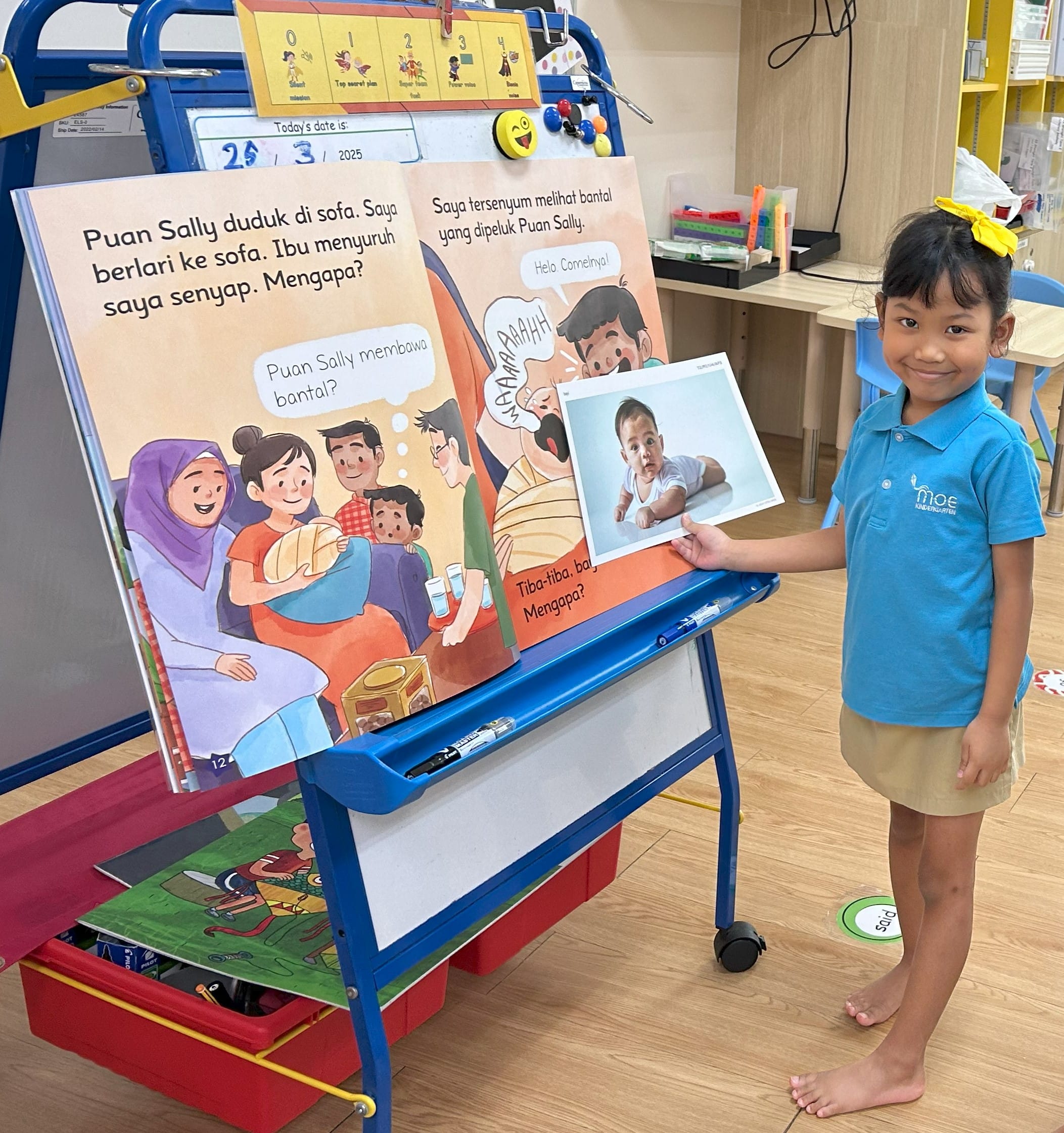 A young girl in a blue MOE Kindergarten uniform holds a baby photo next to a Malay storybook on a classroom easel.