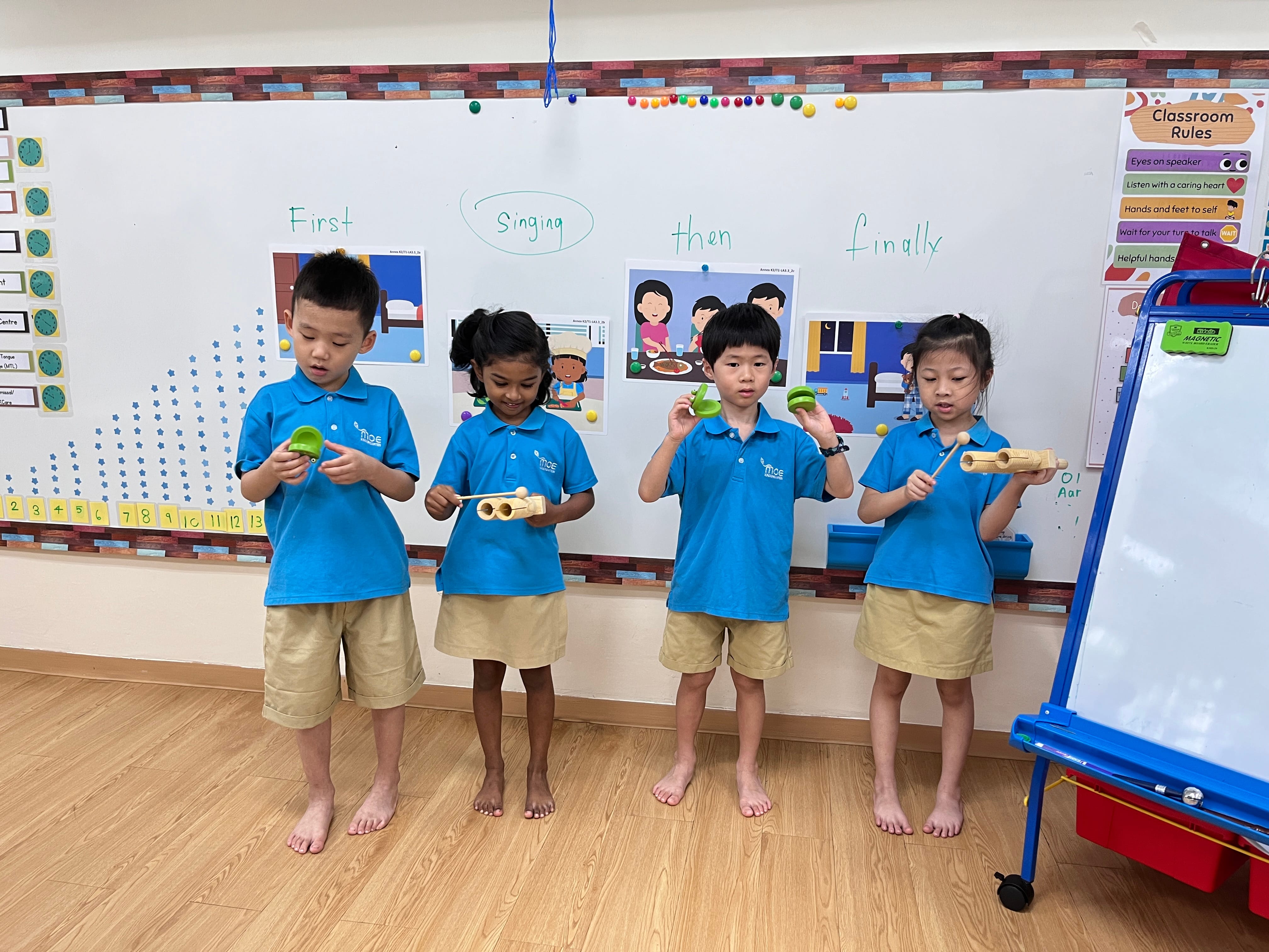 Four young children in blue uniforms stand barefoot holding wooden instruments in front of a classroom whiteboard.