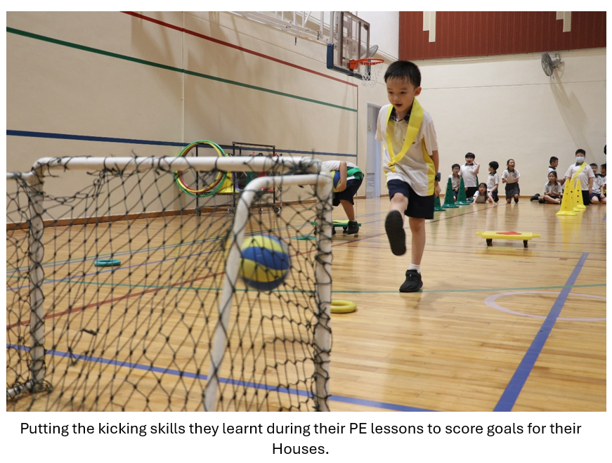 A young boy in a yellow vest kicks a ball into a small goal net in an indoor gymnasium.
