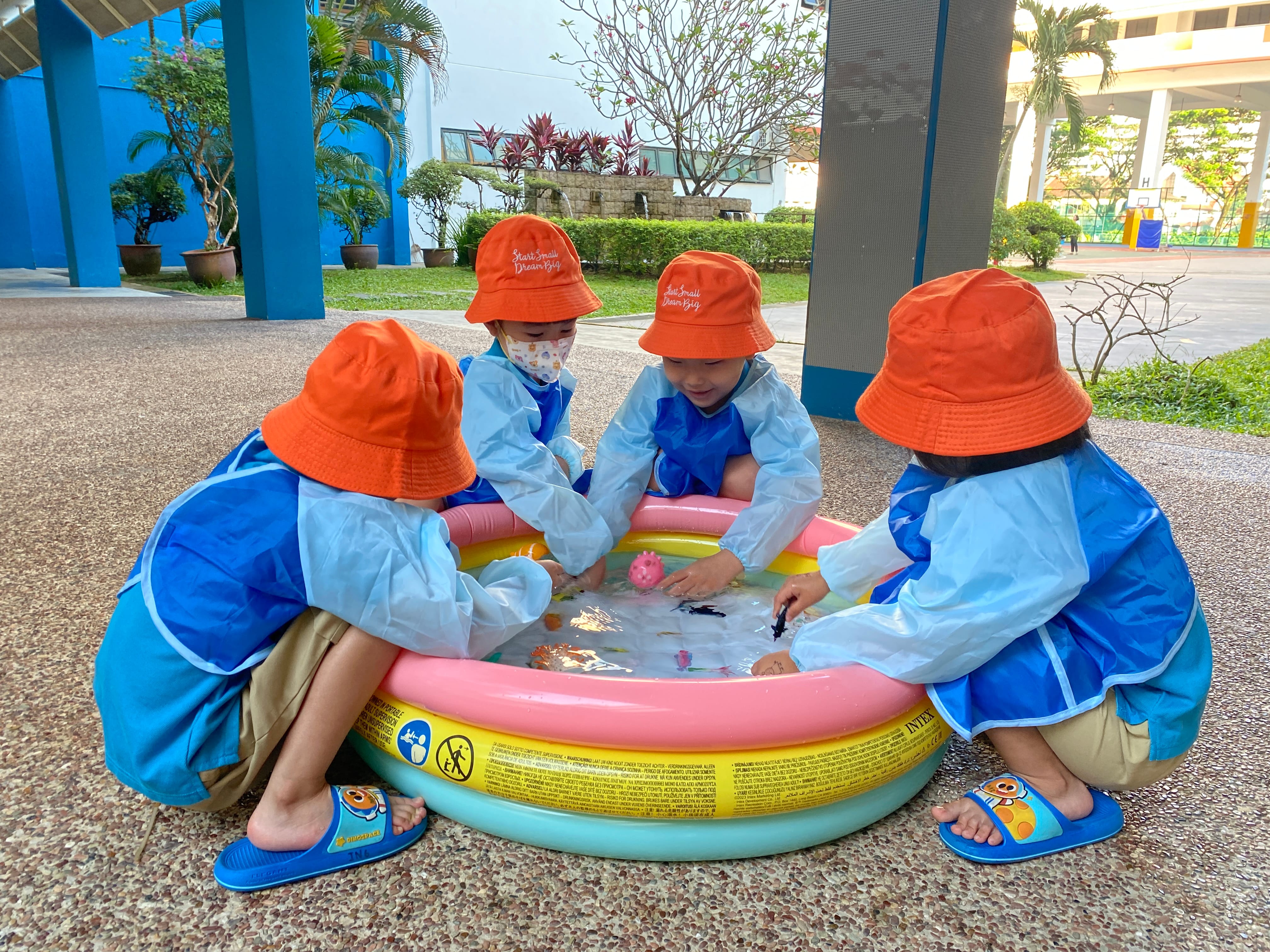 Four young children in orange hats and blue smocks play around a small inflatable pool outdoors.