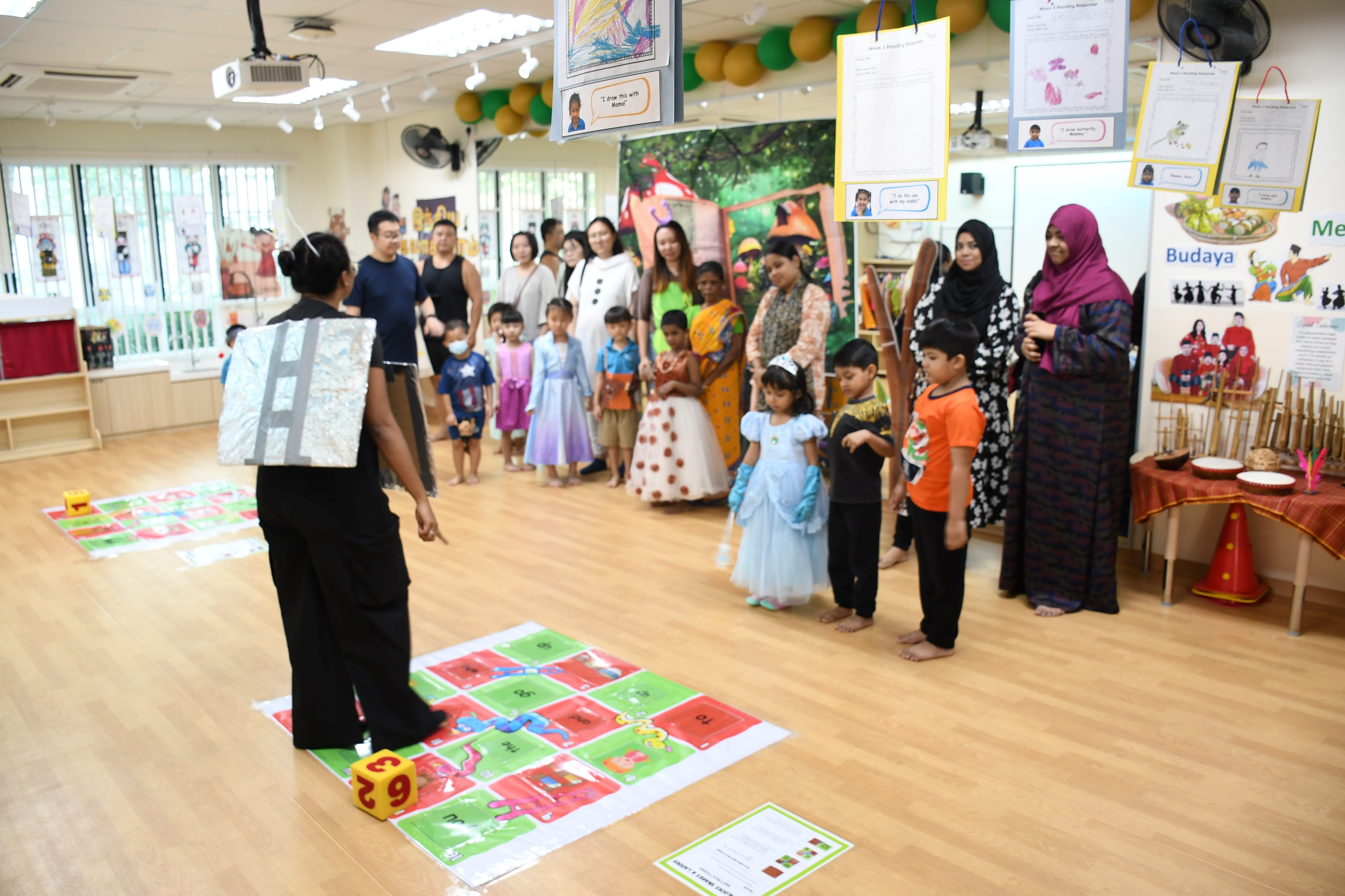 Teacher with a box backpack stands on a floor game mat while children and parents watch in a classroom