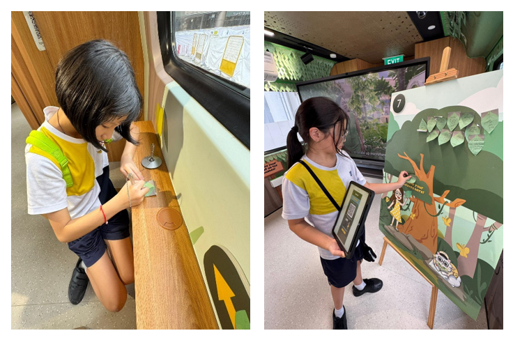 Two children in school uniforms interact with exhibits inside a bus, one kneeling on the floor and one touching a tree display board.