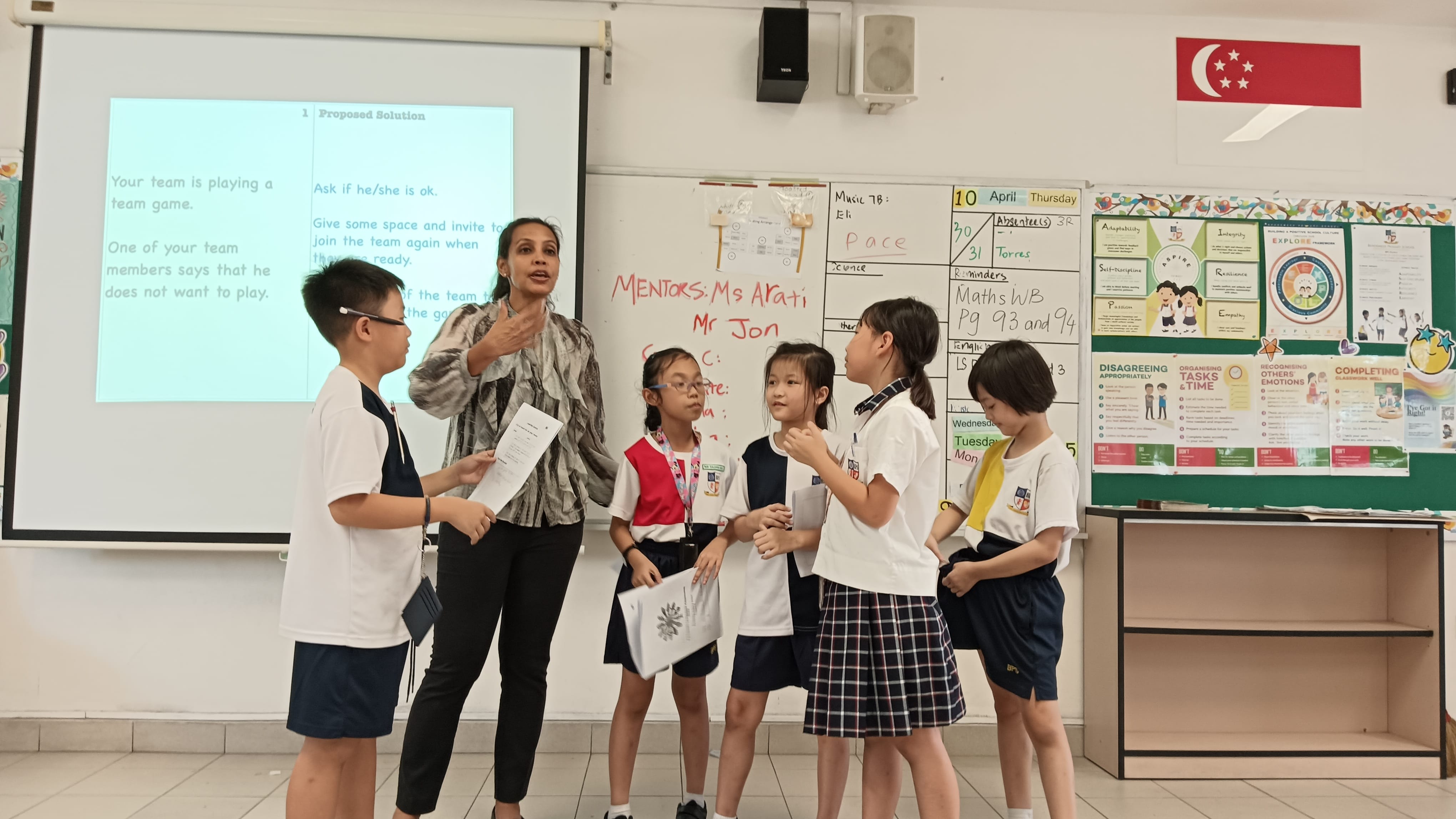 A teacher guides four young students in a classroom activity near a projected slide and whiteboard.