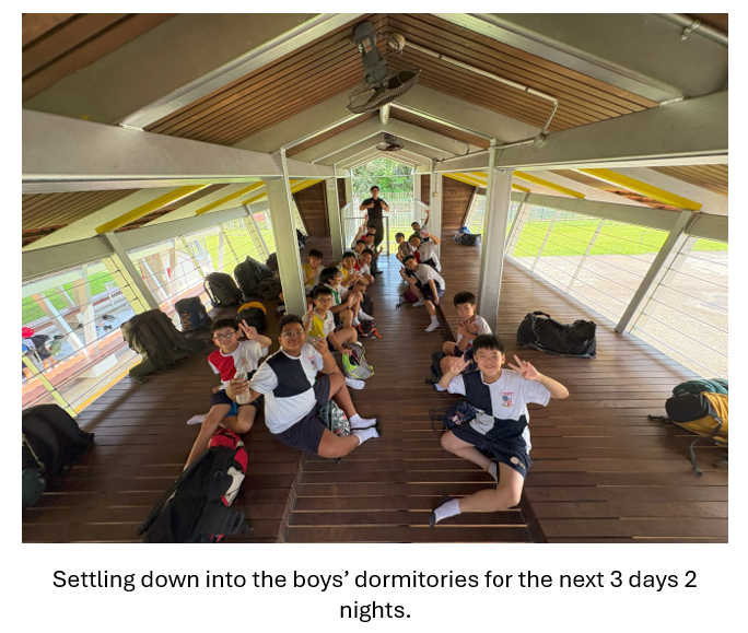 A group of boys sit on a wooden floor in an open-sided pavilion, posing with peace signs and bags nearby.