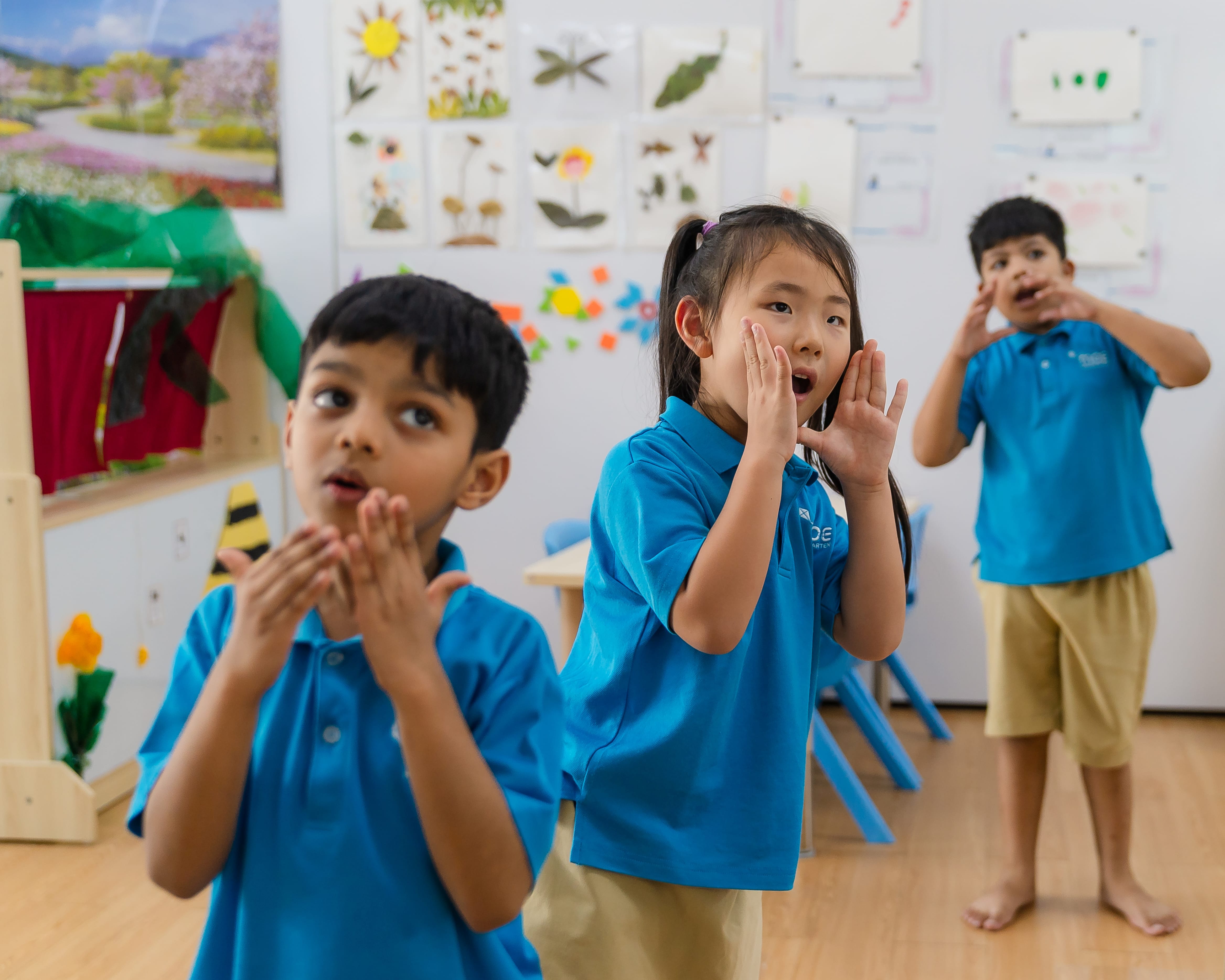 Three children in blue uniforms in a classroom, hands near mouths as if calling or singing.