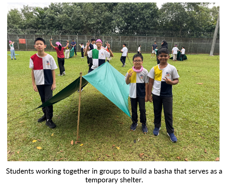 Three students stand proudly next to a small teal tarp shelter on a grassy field with other students in the background.