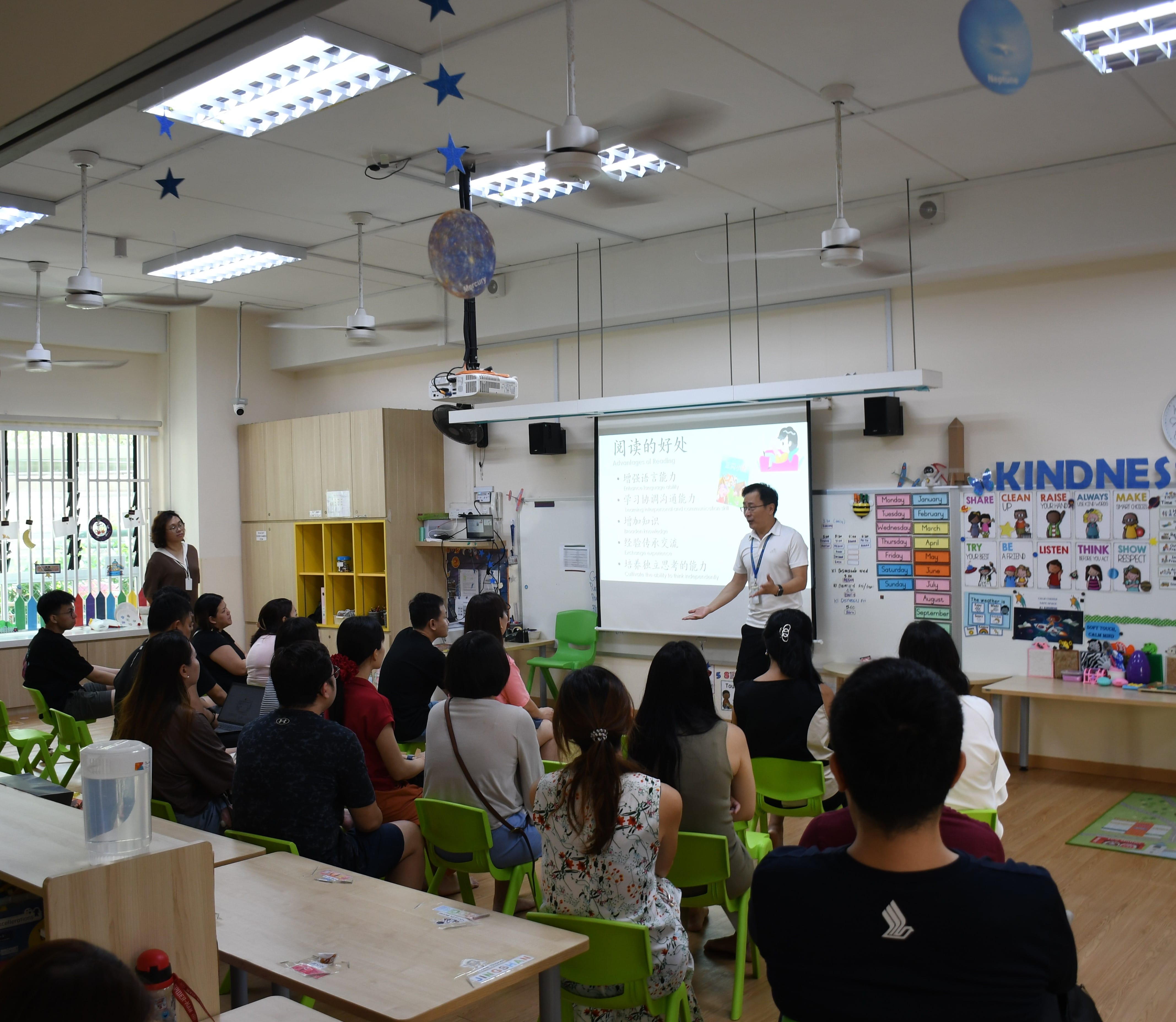 Teacher presents a Chinese-language slideshow to seated adults in a decorated classroom with green chairs.
