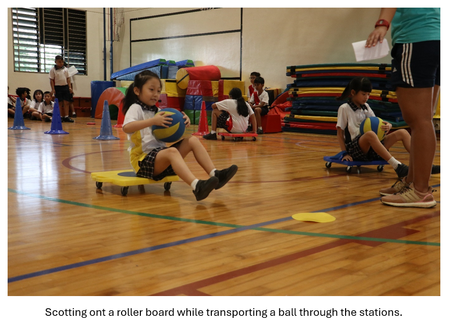 Children in school uniforms sit on wheeled scooter boards in a gym, holding a ball