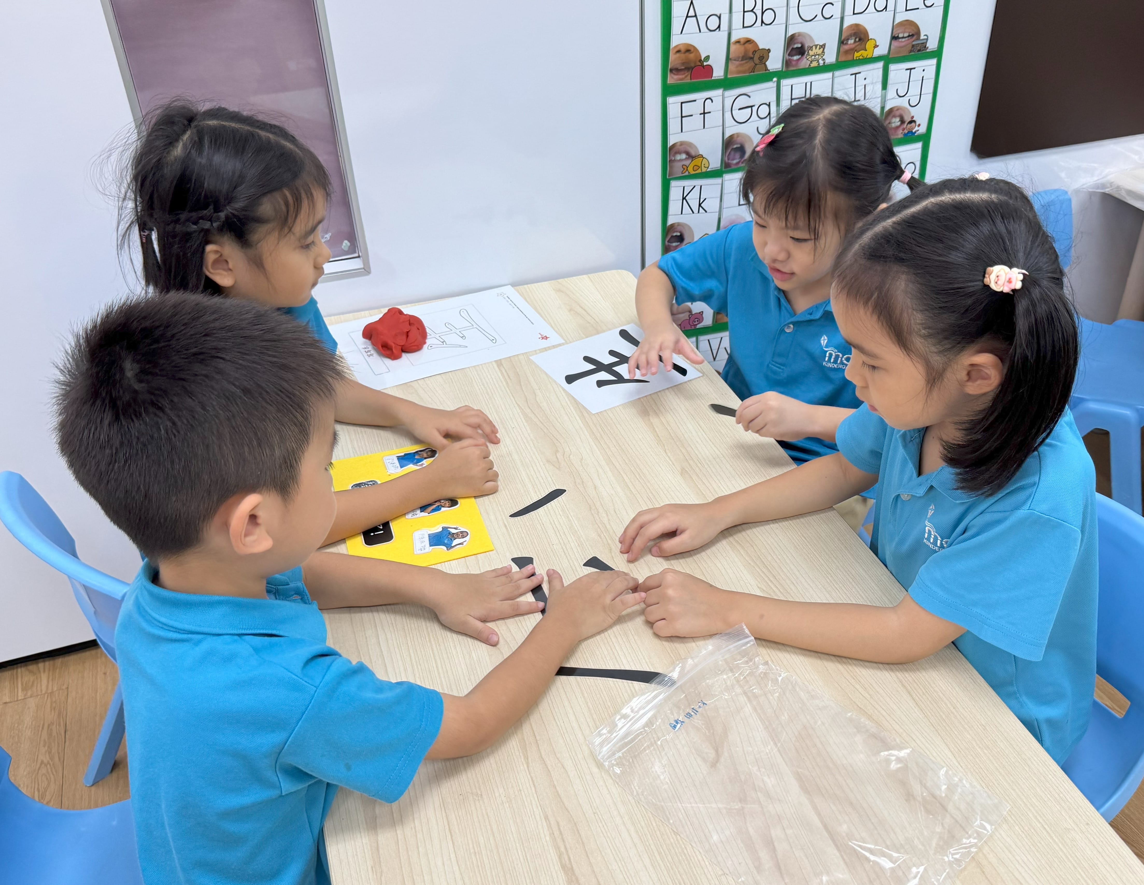 Four young children in blue uniforms sit around a table forming letters with black strips in a classroom.