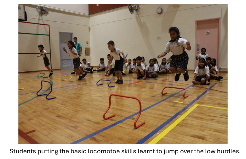 Young students in uniforms jump over colorful low hurdles on a gym floor while classmates watch seated.