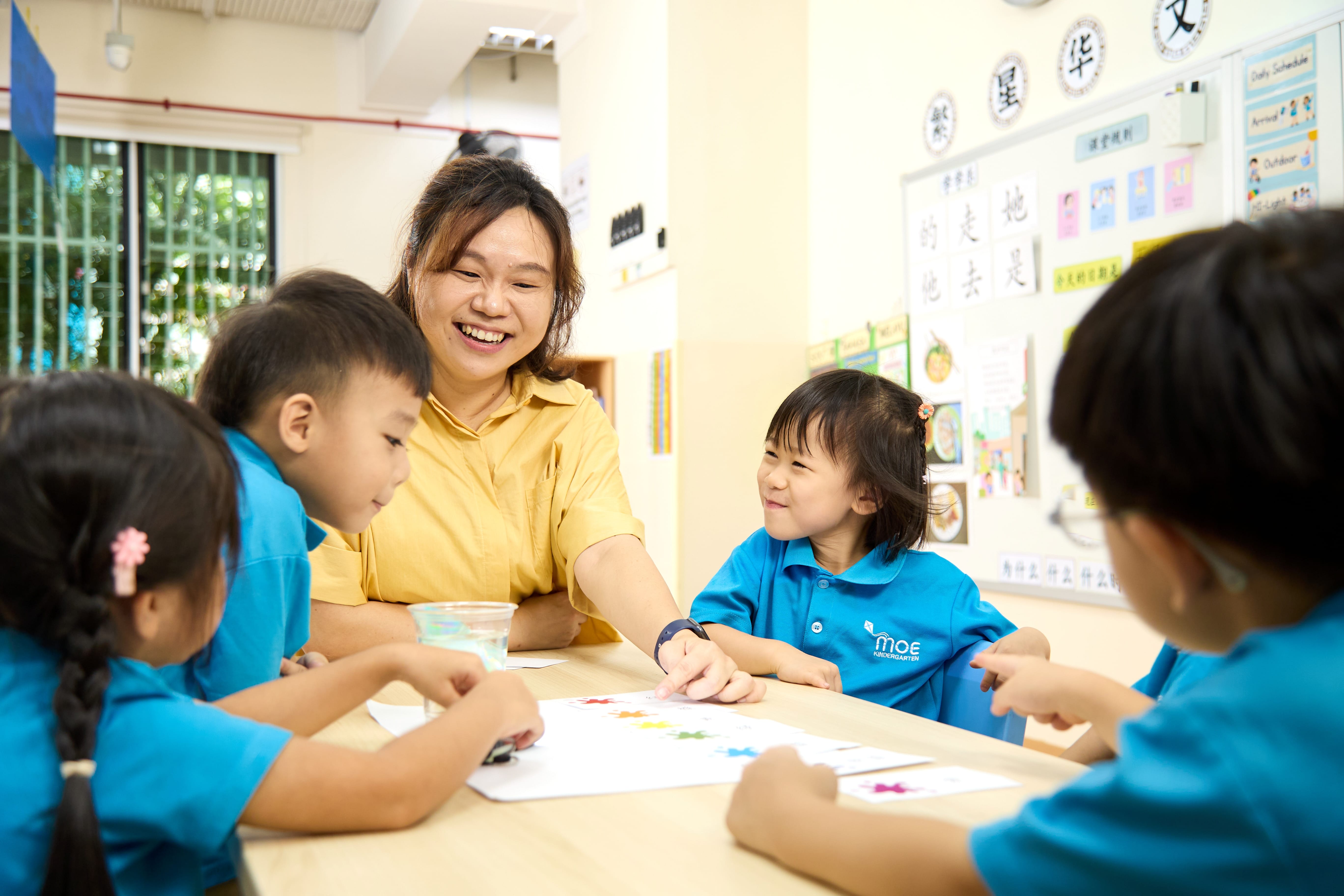 Teacher in yellow smiles with four children in blue uniforms gathered around a table with paper and paint.