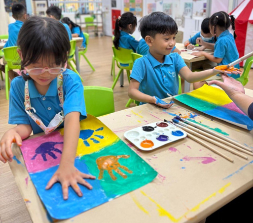 Young children in blue uniforms doing handprint painting with colorful paints on paper in a classroom.