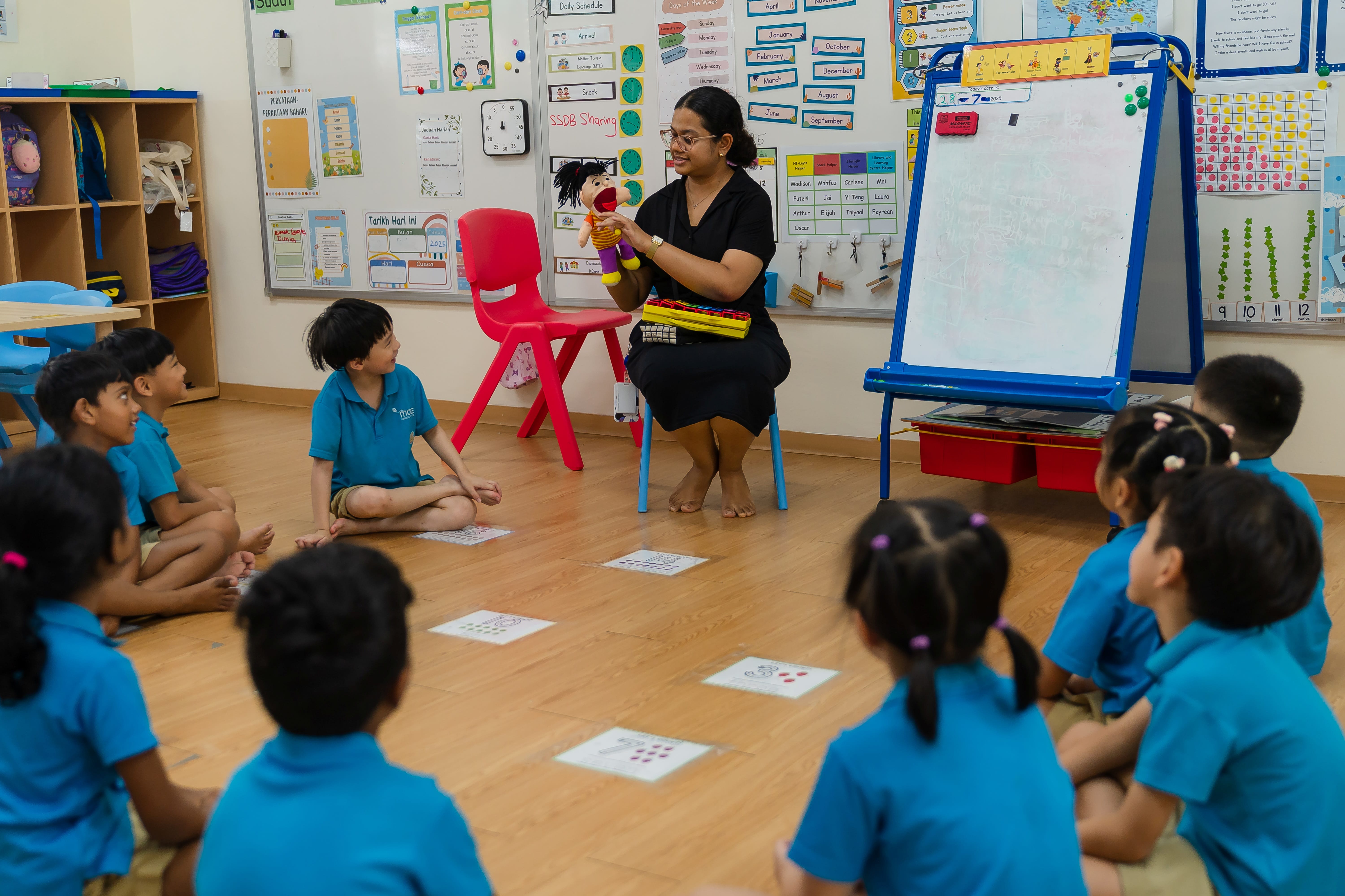 Teacher holds a puppet while young children in blue uniforms sit in a circle on a classroom floor.