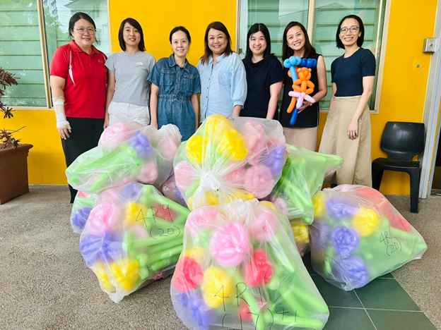 Seven women stand behind large clear bags filled with colorful balloon flower sculptures against a yellow wall.