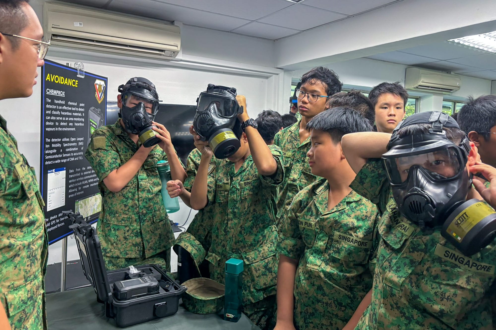 Young Singapore soldiers in camouflage uniforms try on black gas masks indoors near an Avoidance Chemprox display board.