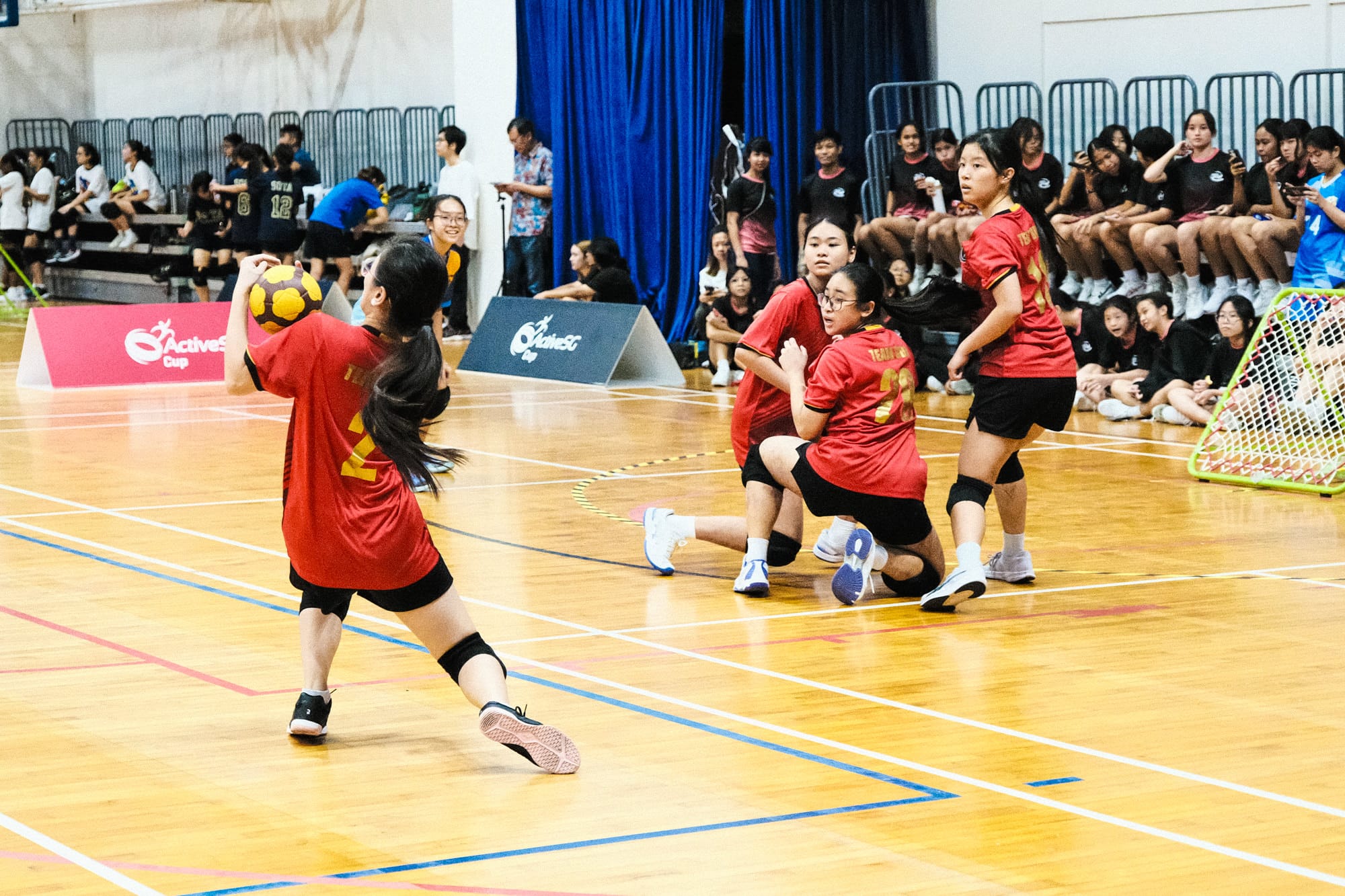 Girls in red jerseys play indoor handball on a wood court, watched by seated spectators along the wall.