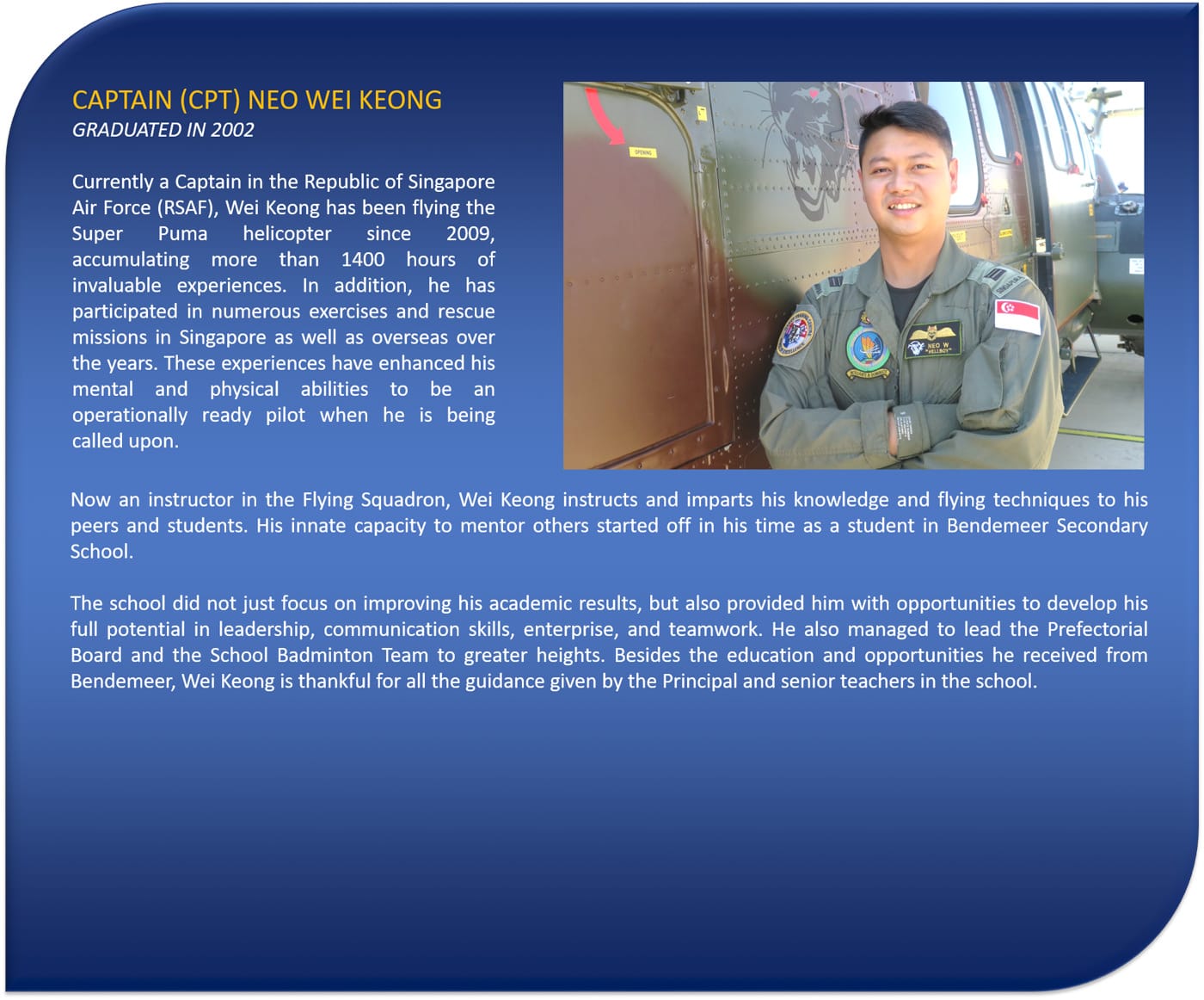 A smiling man in a green flight suit with patches stands arms crossed beside a military helicopter.