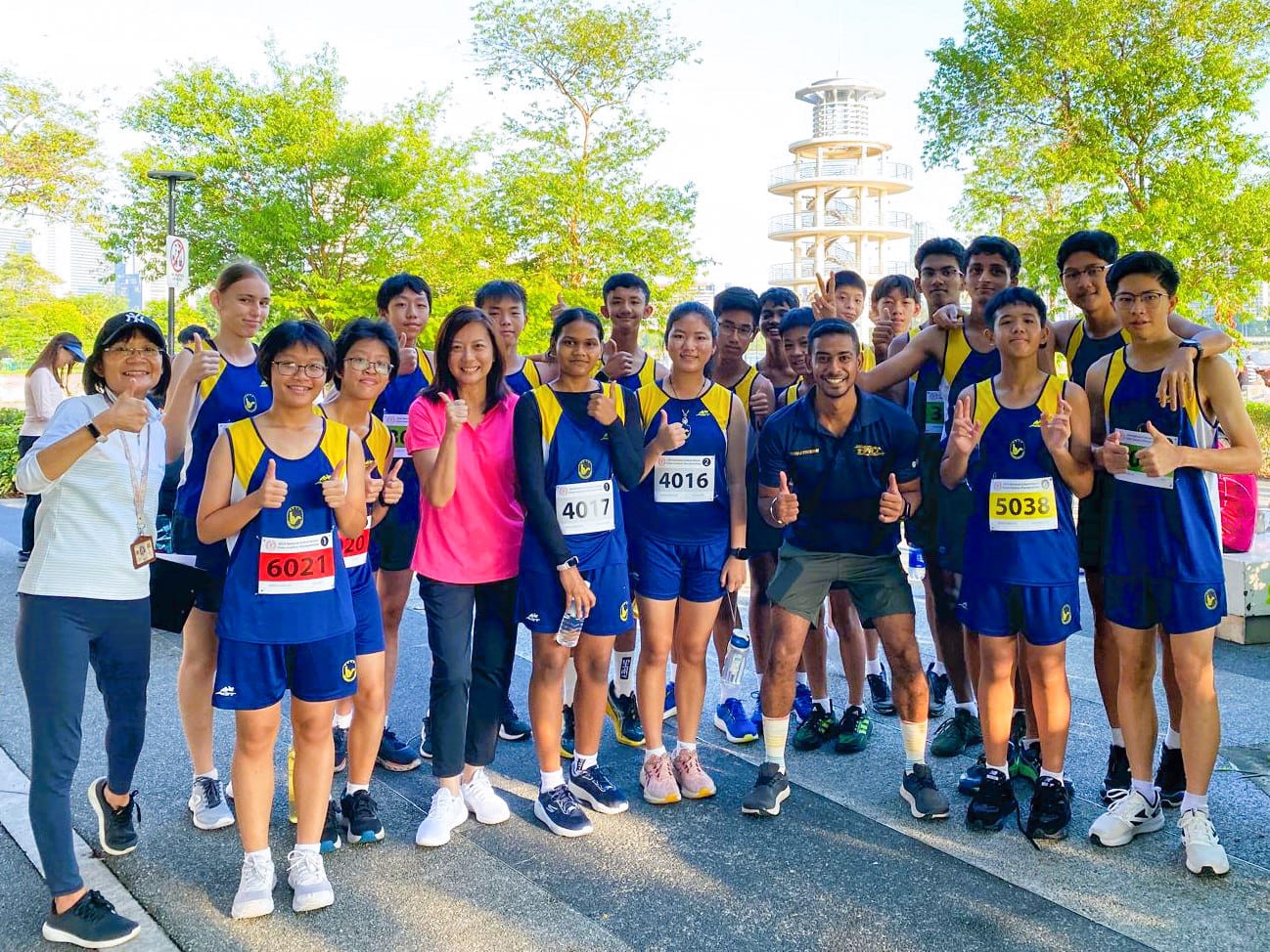 A group of young runners in blue and yellow uniforms with race bibs pose with thumbs up outdoors near a tower.