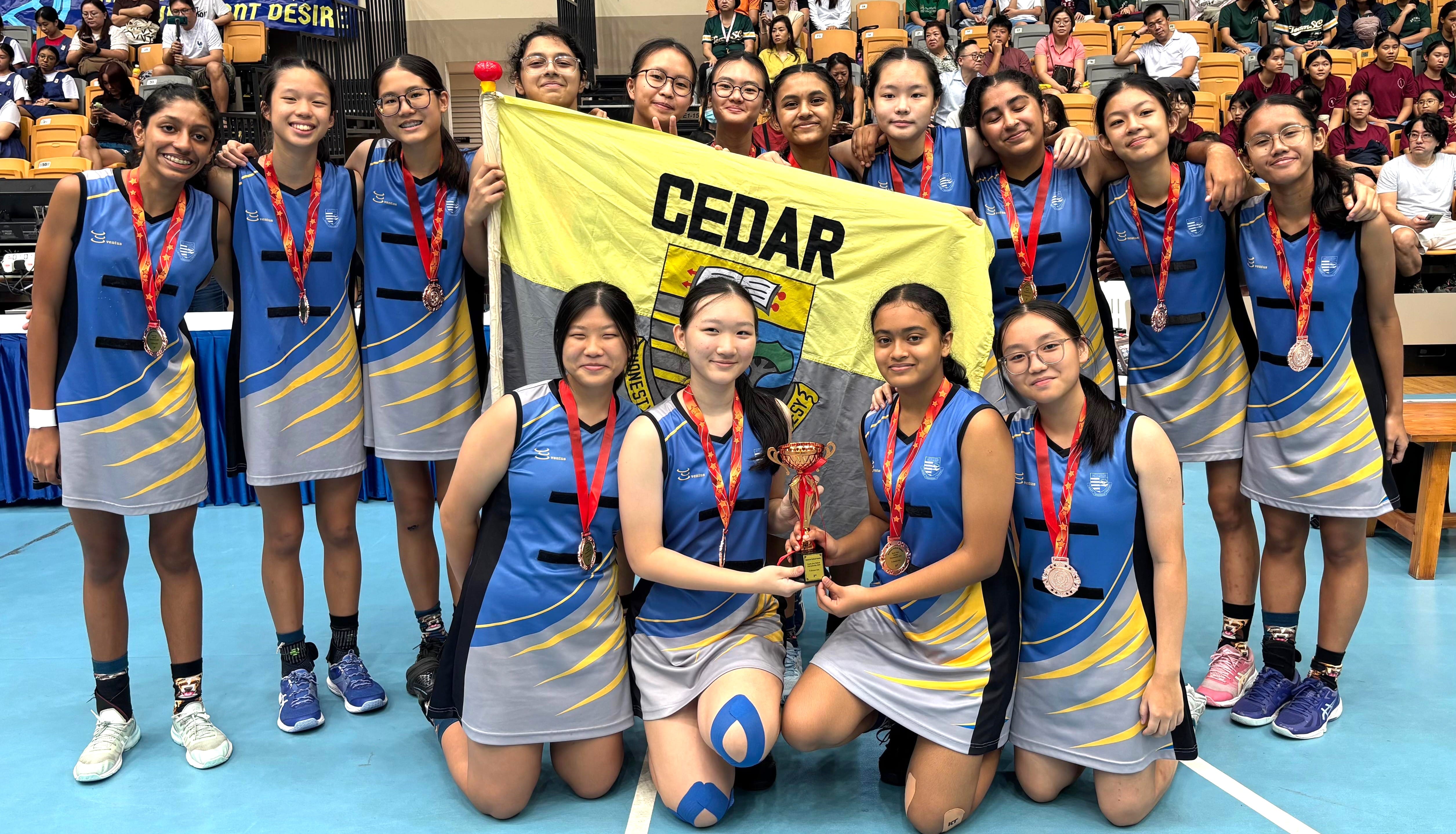 Cedar school girls sports team posing with medals, a trophy, and a yellow Cedar flag in an indoor court.