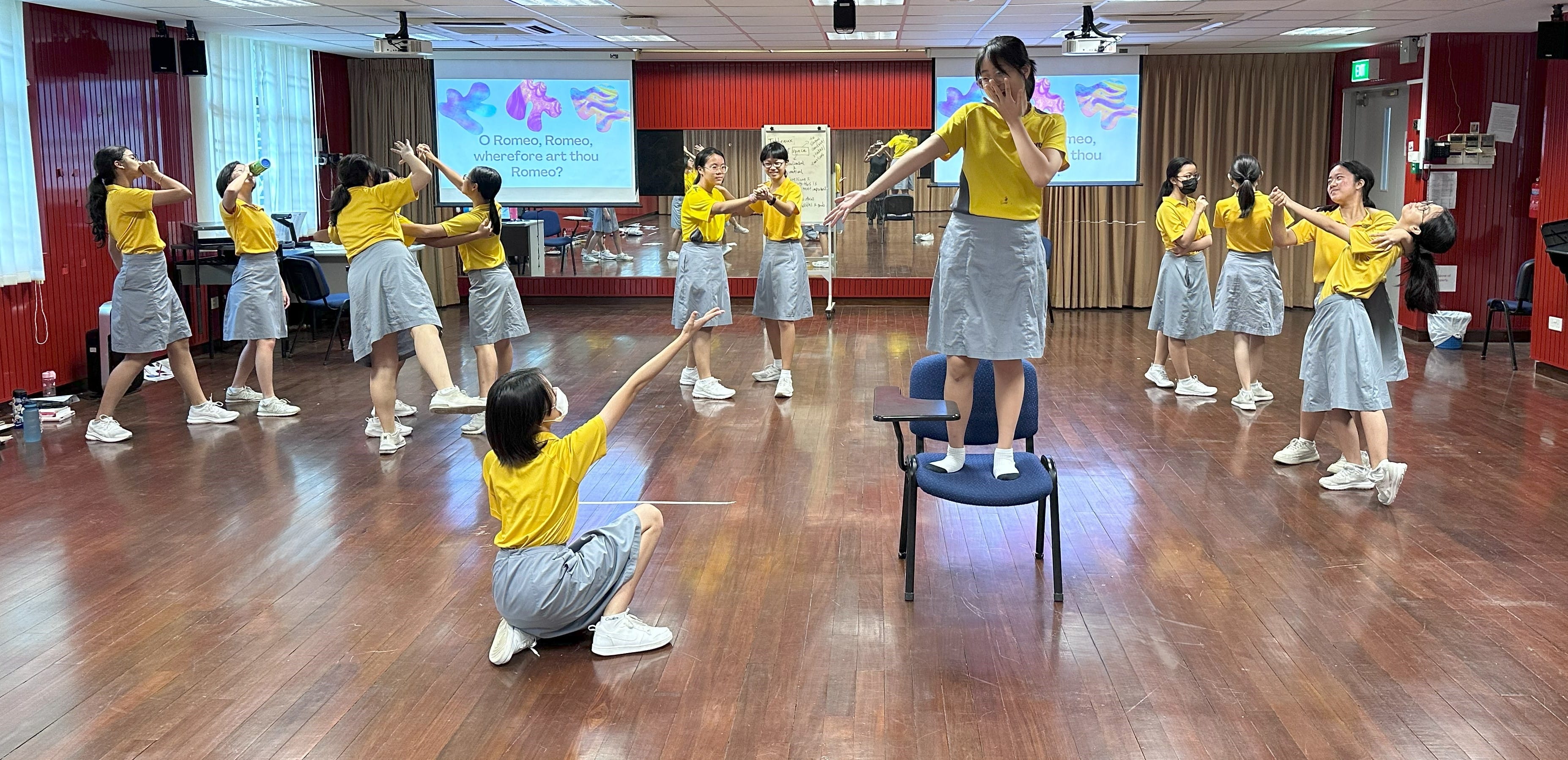 Students in yellow tops and grey skirts act out Romeo and Juliet in a hall with projected text on screens.