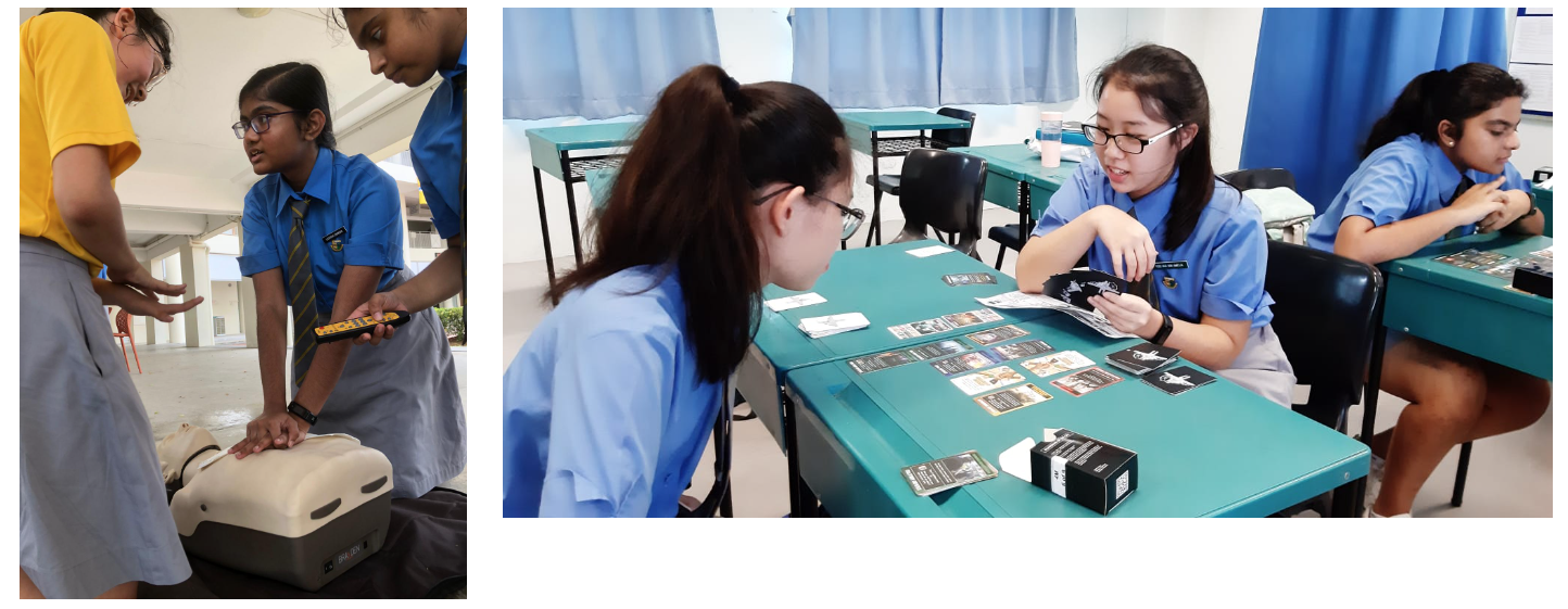 Students in blue uniforms practice CPR on a mannequin and review cards at a teal desk in a classroom.