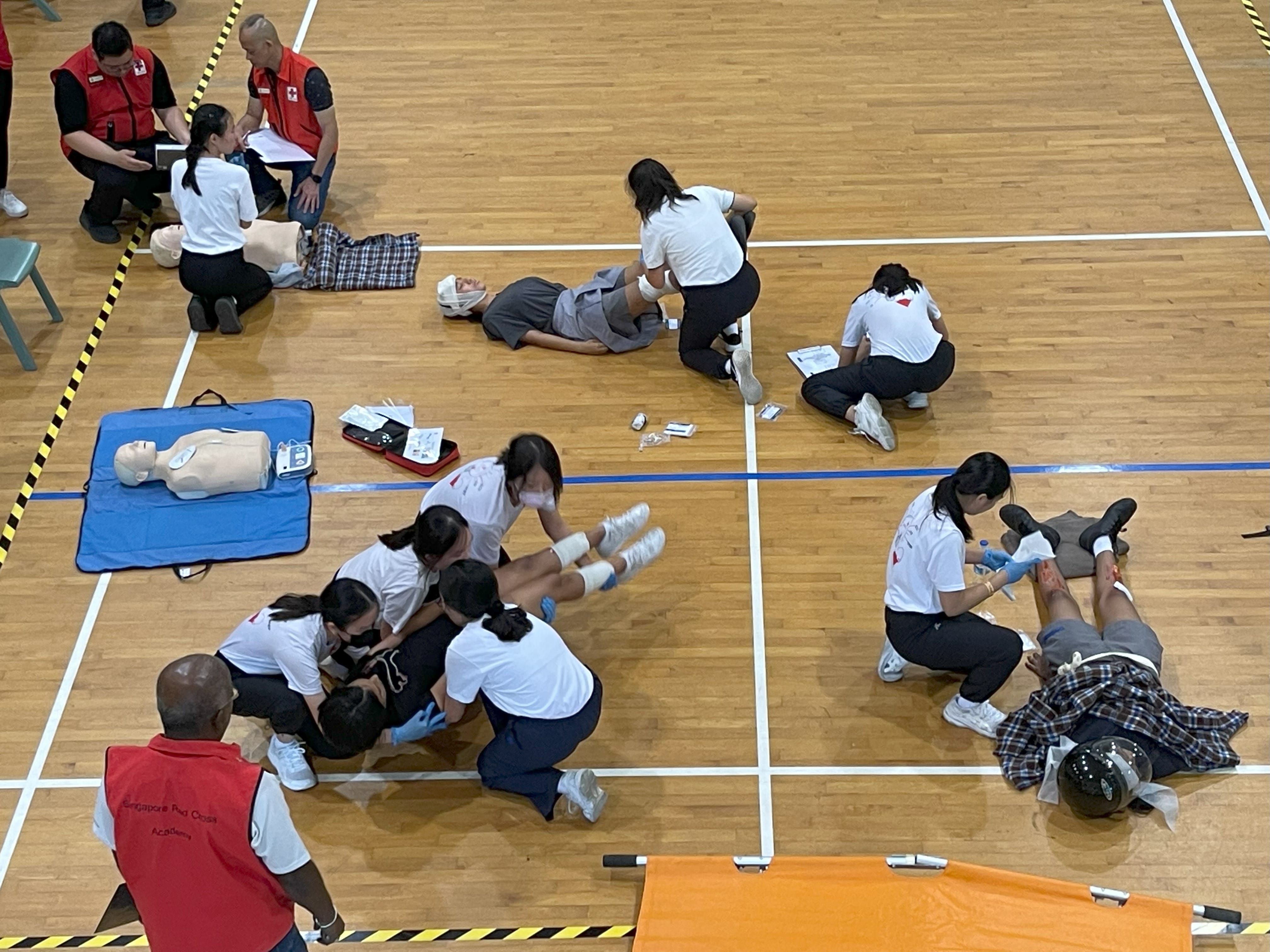 Students in white shirts practice first aid on mannequins and role-play patients on a gym floor, supervised by Red Cross instructors.