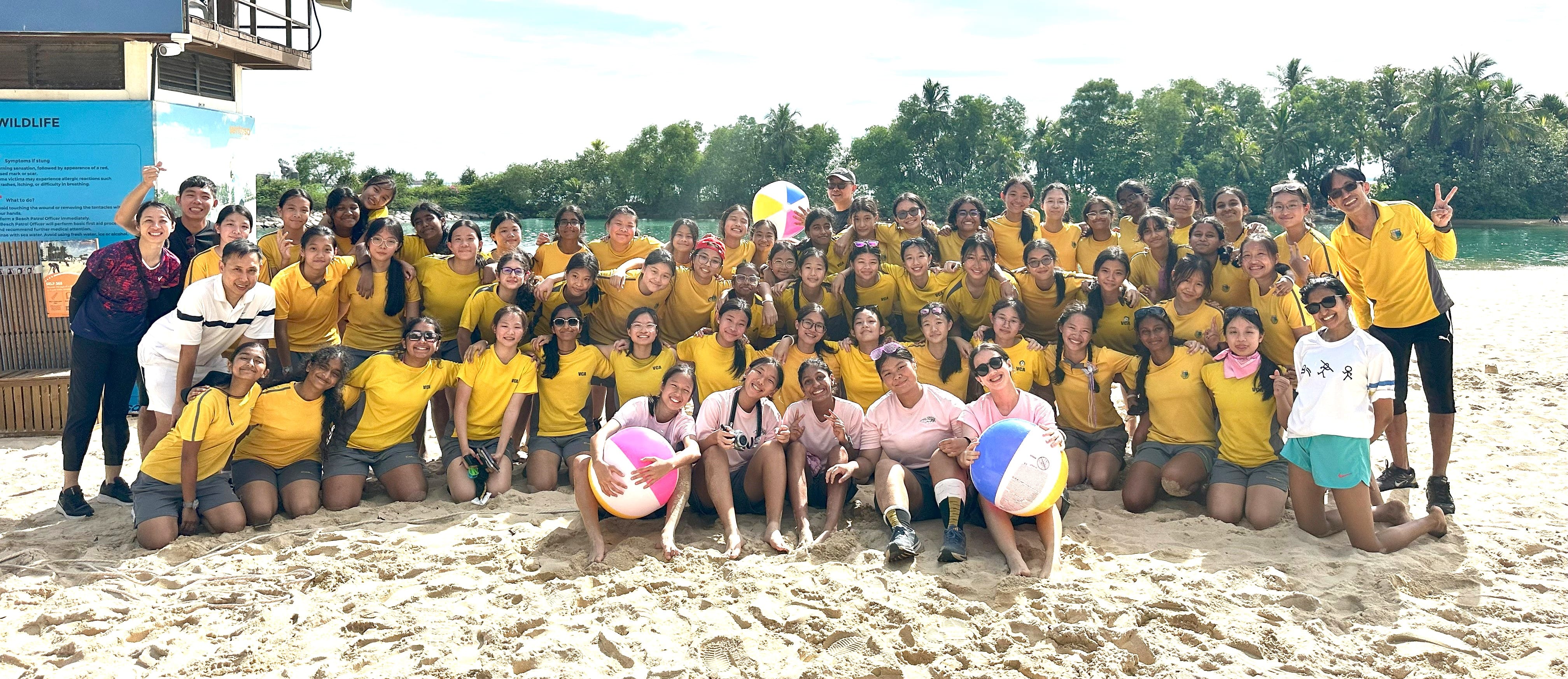 Large group of students in yellow shirts posing together on a sandy beach, some holding colorful beach balls.